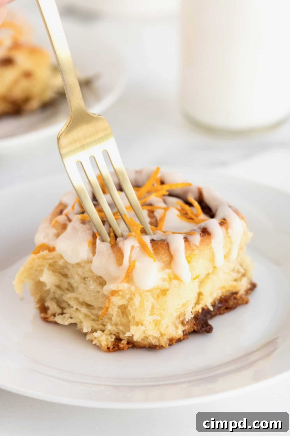 A fork gently pressing into a warm cinnamon roll, topped with vibrant orange zest, resting on a clean white plate, highlighting its tender texture.
