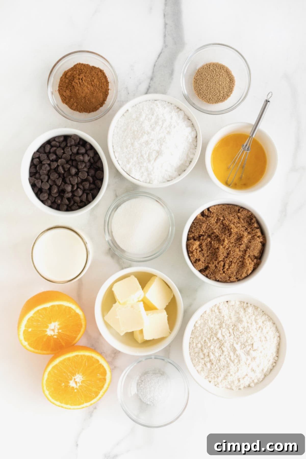 Various ingredients for Chocolate Orange Cinnamon Rolls meticulously arranged in elegant white and glass dishes on a pristine white marble counter.