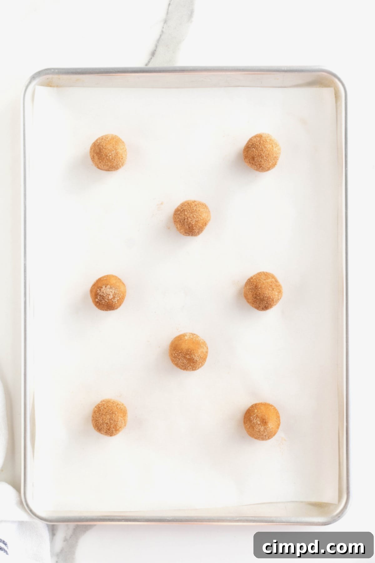 Eight evenly spaced gingerdoodle cookie dough balls, coated in spice sugar, arranged on a parchment-lined aluminum baking sheet, ready for the oven.