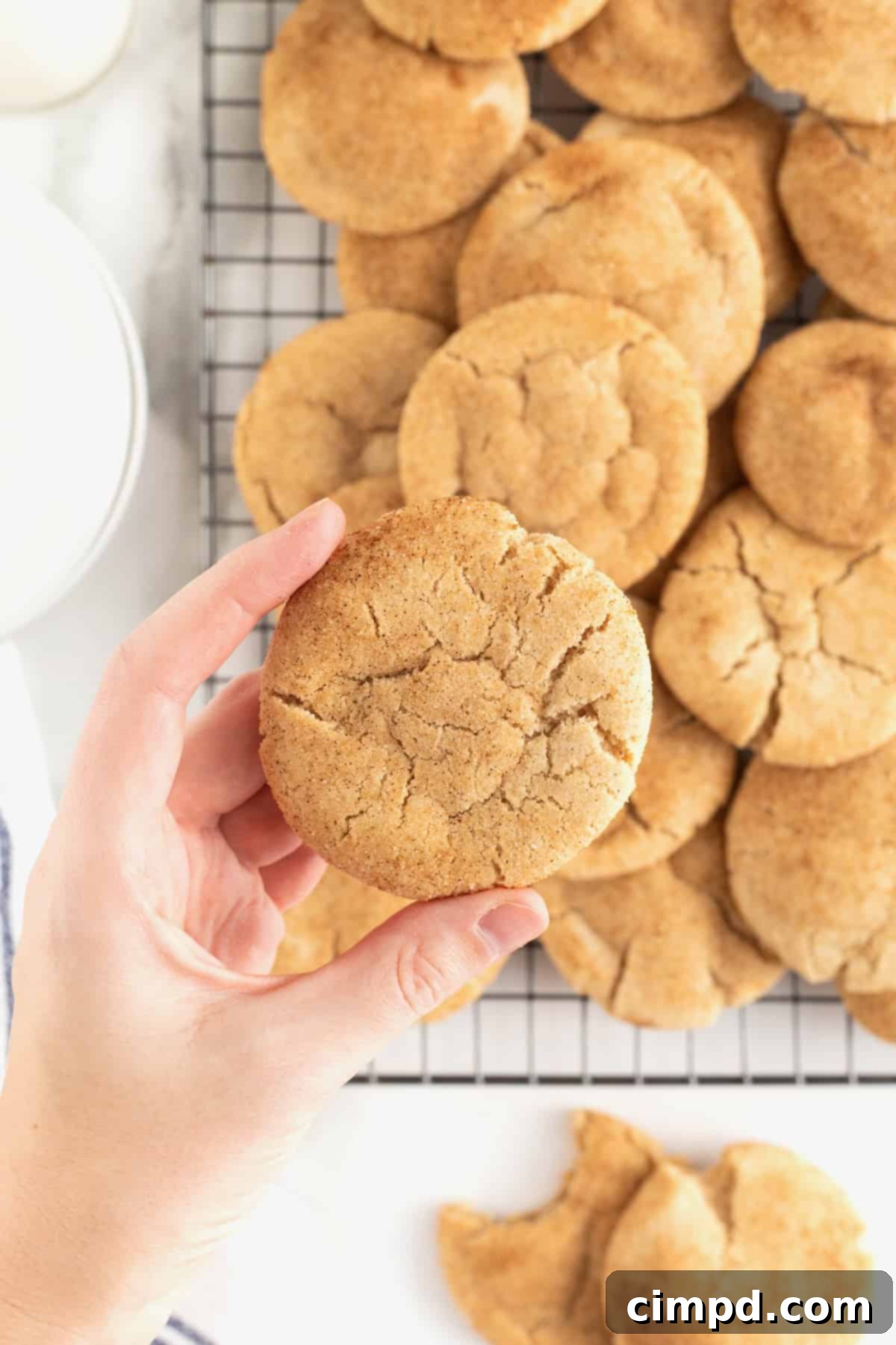 A hand gently lifts a warm gingerdoodle cookie from a wire cooling rack, which is filled with more freshly baked gingerdoodle cookies.