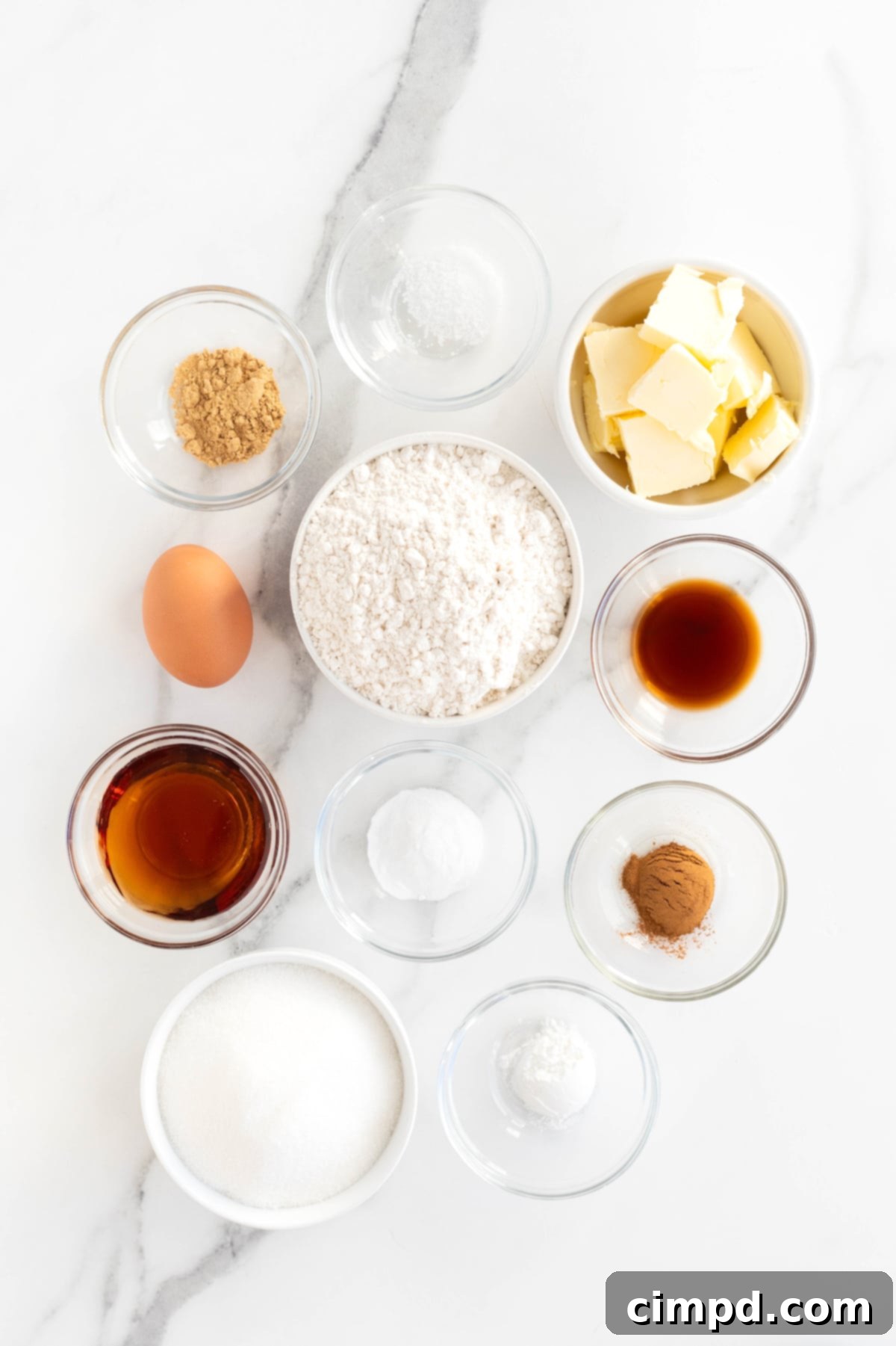 A selection of raw ingredients for gingerdoodle cookies, including flour, sugar, butter, and various spices, neatly arranged in small glass containers on a white marble counter.