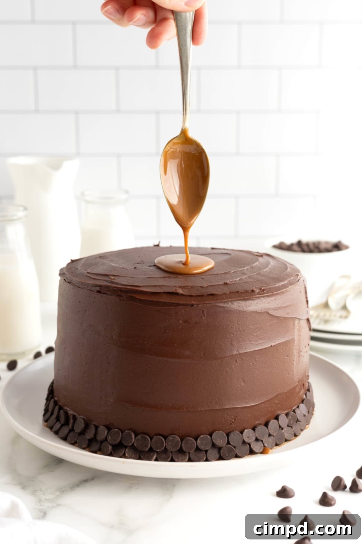 A metal spoon, generously coated in luscious caramel, held vertically over the top of a chocolate layer cake, poised for the final drizzle.