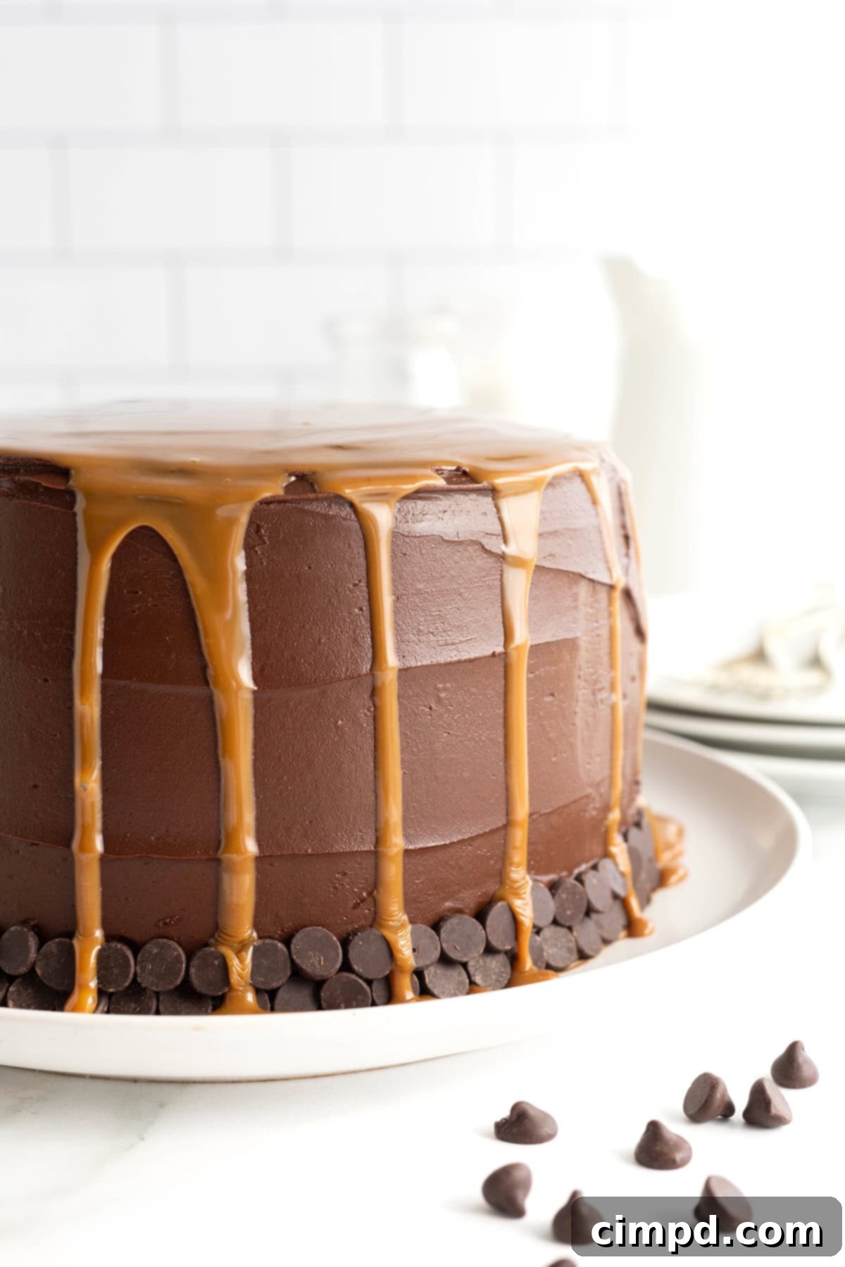 A stunning three-layer chocolate cake with a lavish caramel drizzle flowing elegantly down its sides, presented on a pristine white cake plate.