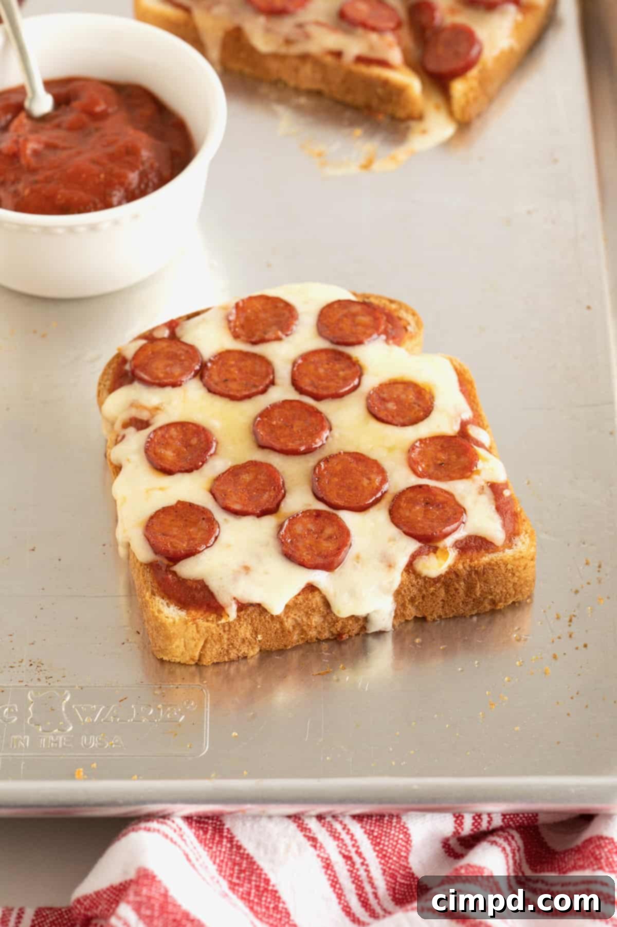 Crispy pizza toast adorned with mini pepperonis, resting on a baking sheet, with a small bowl of marinara sauce nearby.