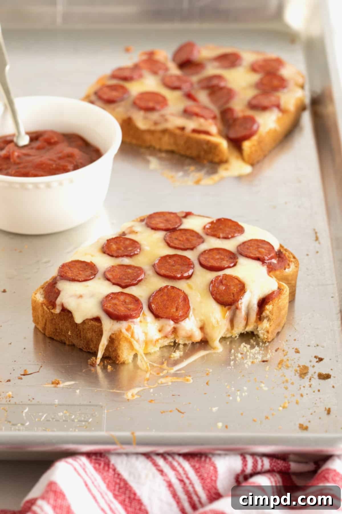 A slice of pizza toast, with a bite taken, on an aluminum baking sheet, accompanied by a small white dish of marinara sauce.