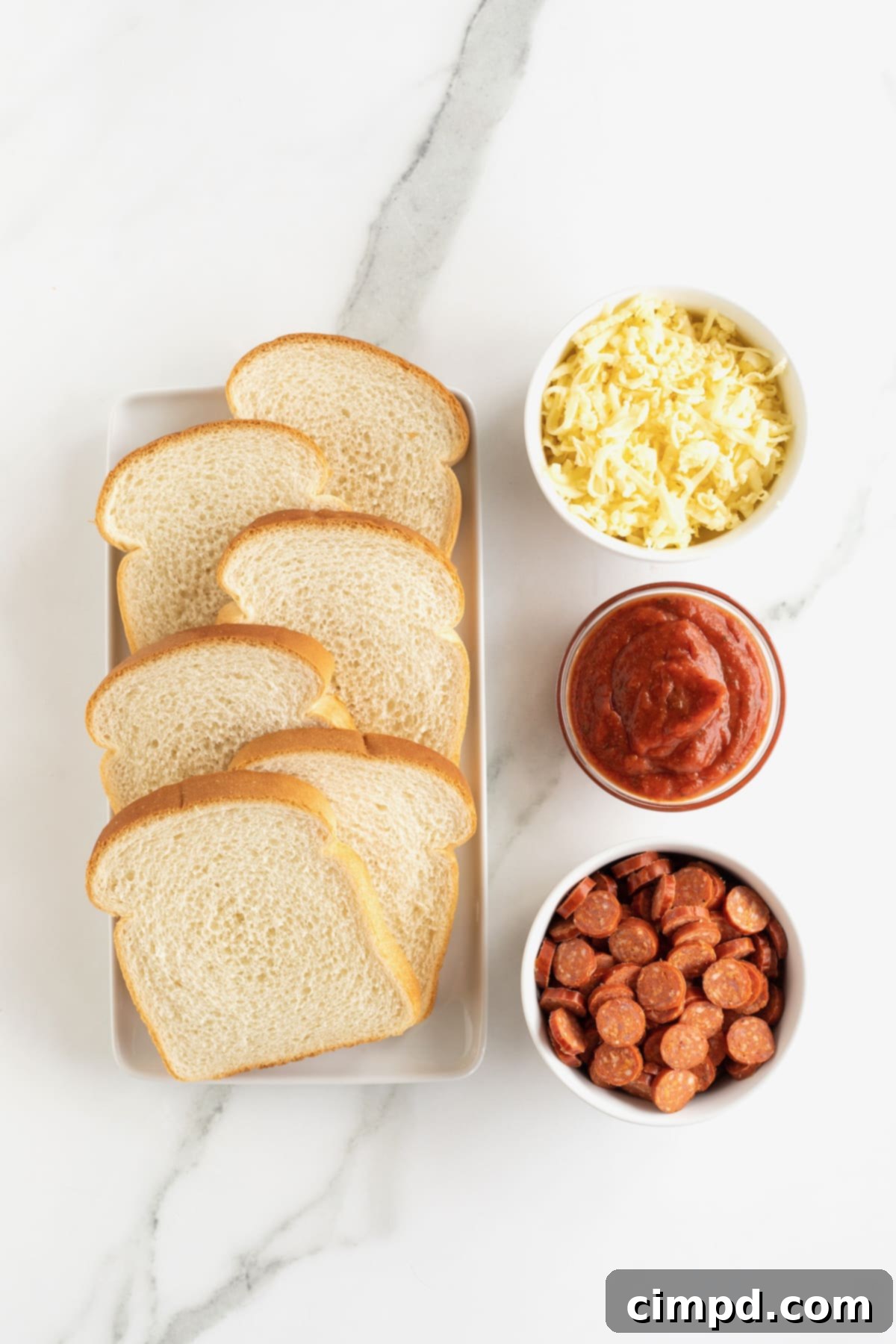 Arrangement of white bread, shredded mozzarella cheese, mini pepperonis, and pizza sauce in white containers on a pristine white marble counter, ready for pizza toast preparation.