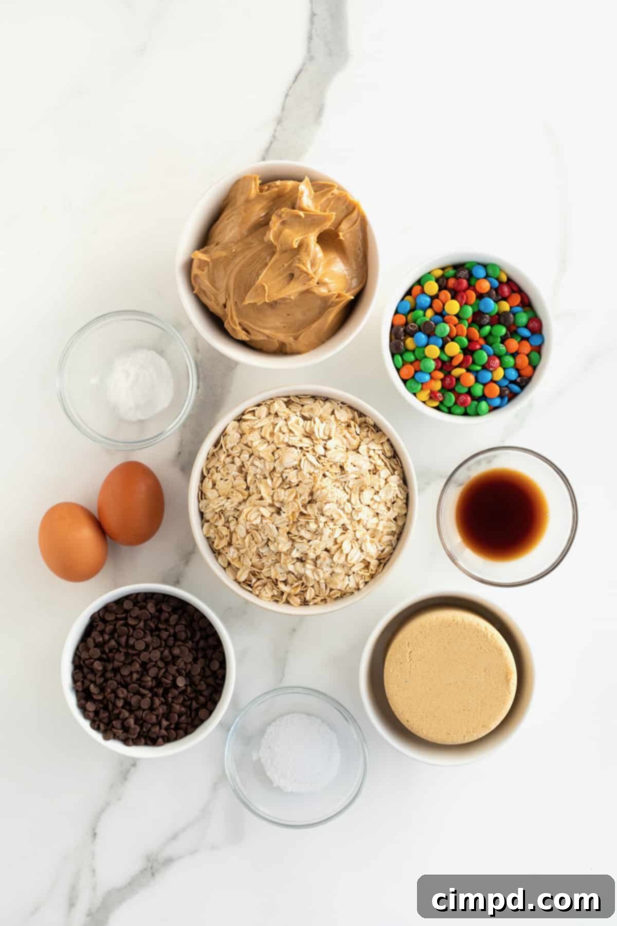 An array of ingredients for monster cookie bars, meticulously arranged in clear glass dishes on a pristine white marble counter, ready for baking.