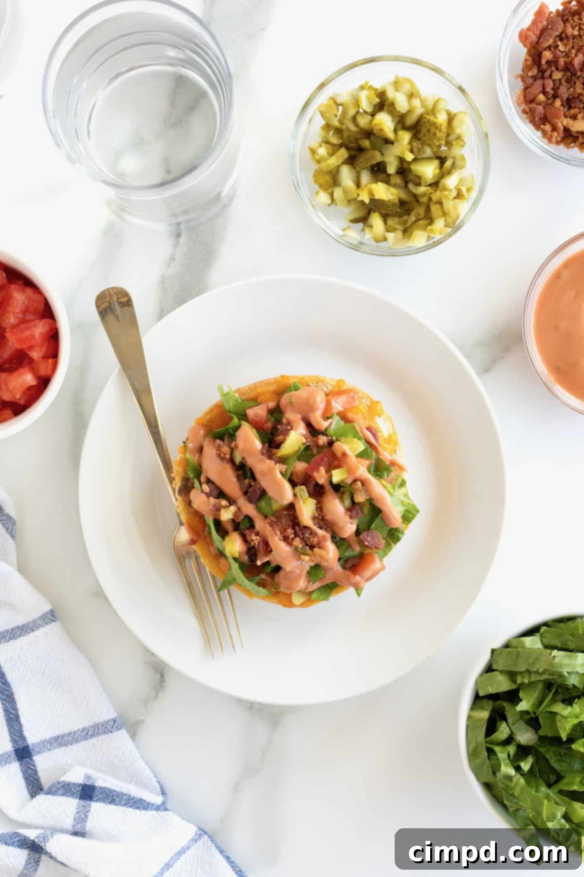 A single cheeseburger bun pizza on a small white plate with a fork, showing the layers of beef, cheese, and fresh toppings.