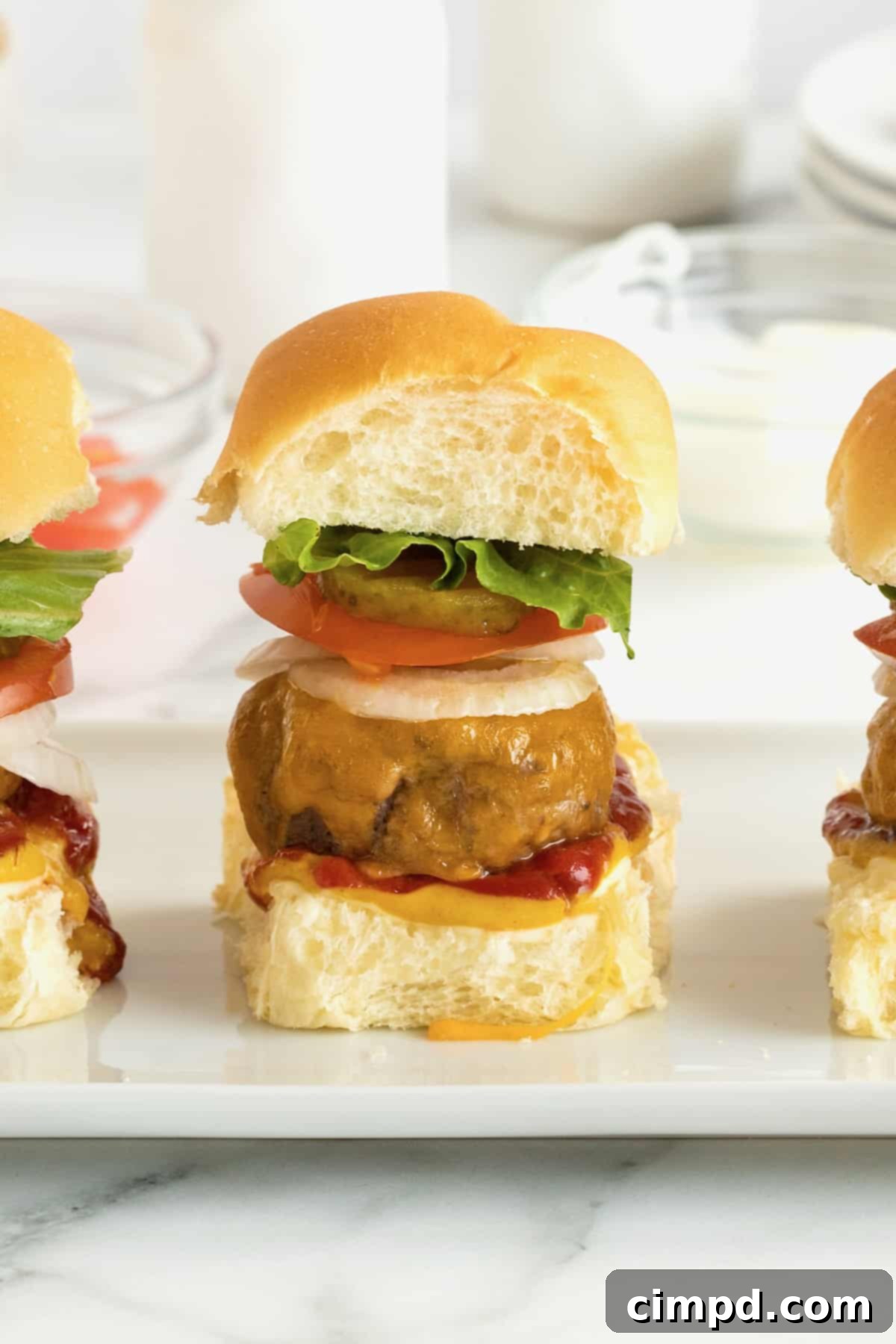 Three mini cheeseburgers garnished with onion, tomato, pickle, and lettuce on a pristine white serving plate.