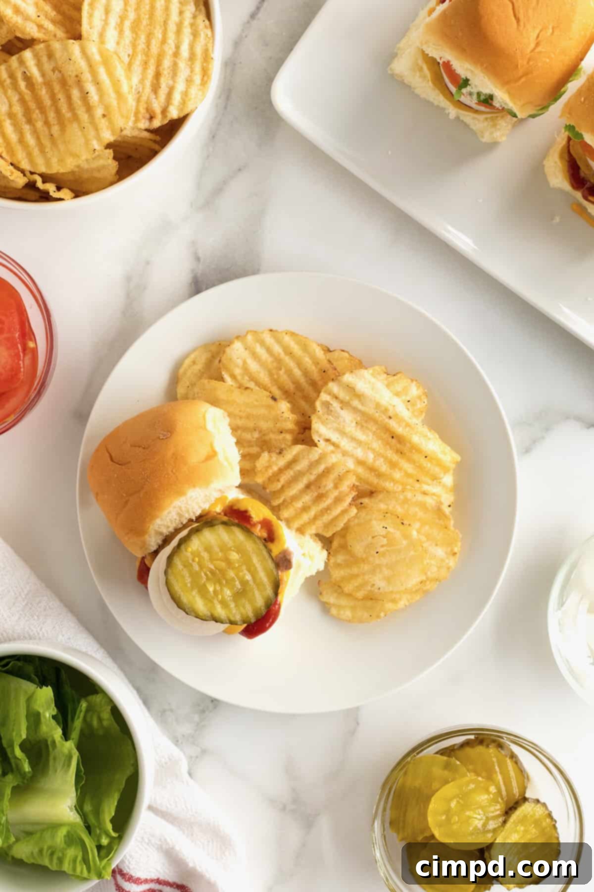 A white dinner plate holding a skillet slider with the top bun removed, revealing a crisp pickle slice and a side of wavy cut potato chips.