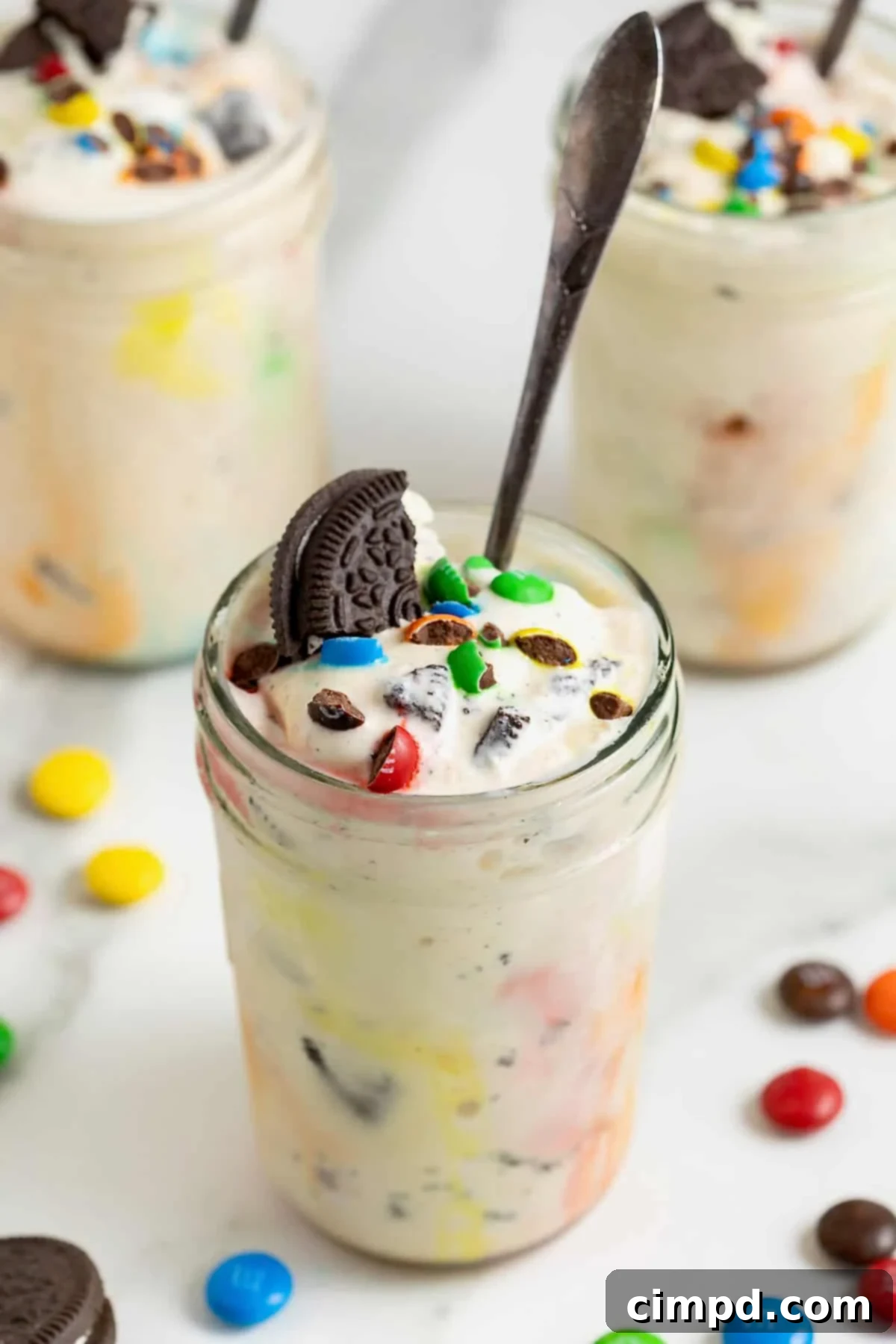 A homemade M&M Blizzard garnished with a piece of Oreo in a wide mouth jar. There is a spoon sticking out of the jar, ready to be enjoyed.