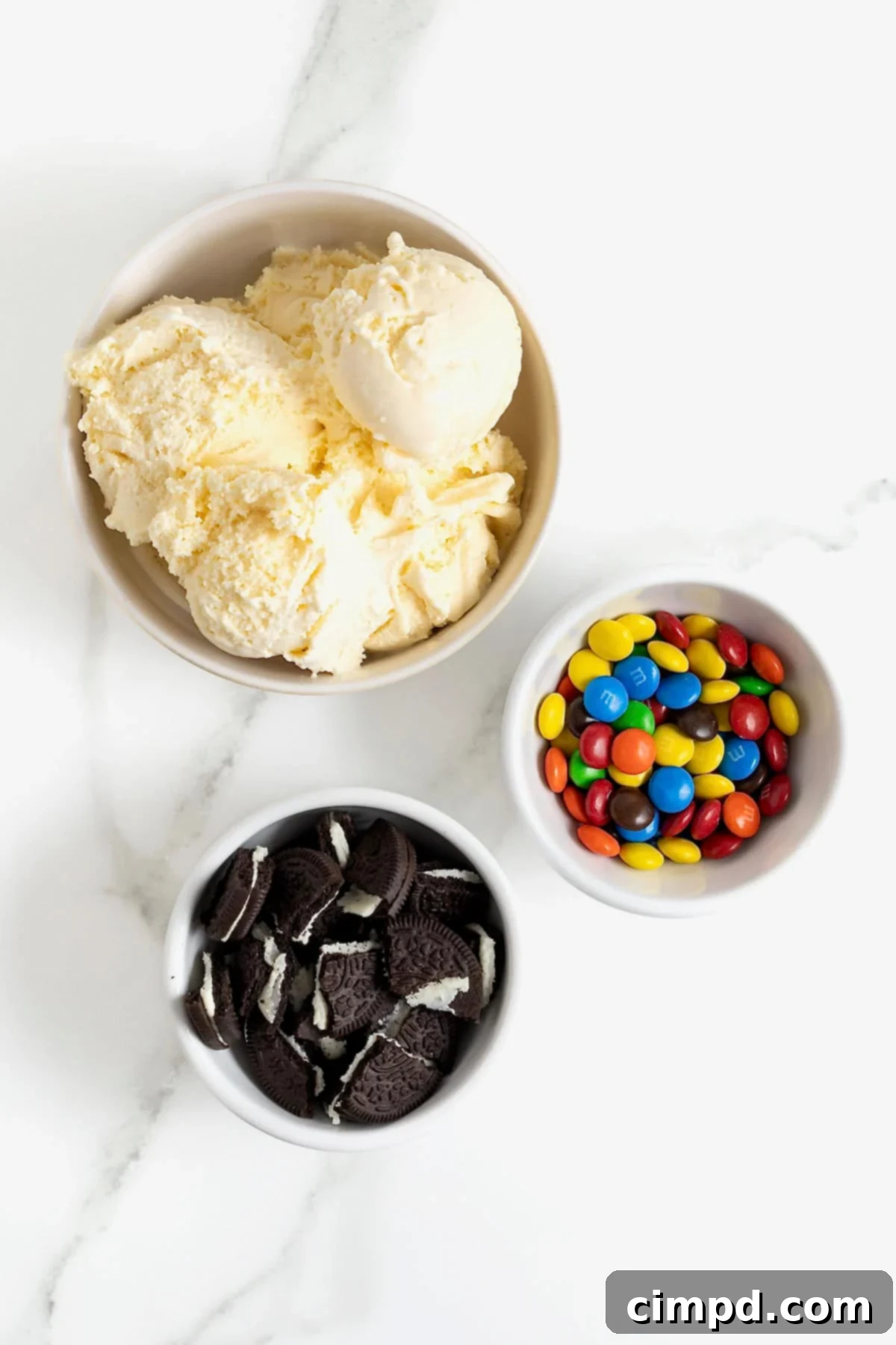 Ingredients for a homemade Oreo and mini M&M blizzard, including vanilla ice cream, crushed Oreos, and mini M&Ms, arranged in white dishes on a white marble counter, ready for mixing.
