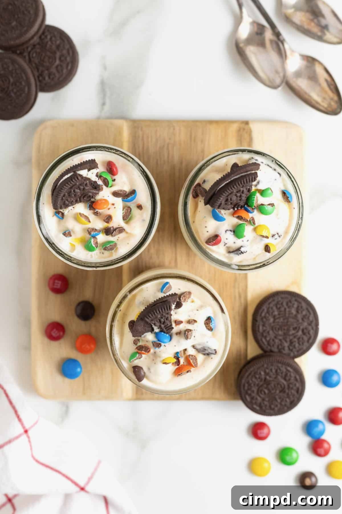 Three homemade blizzards in mason jars, topped with various mix-ins like Oreos, M&Ms, and sprinkles, beautifully presented on a square light wood cutting board.