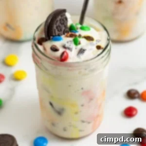 A homemade M&M Blizzard garnished with a piece of Oreo in a wide mouth jar. There is a spoon sticking out of the jar.