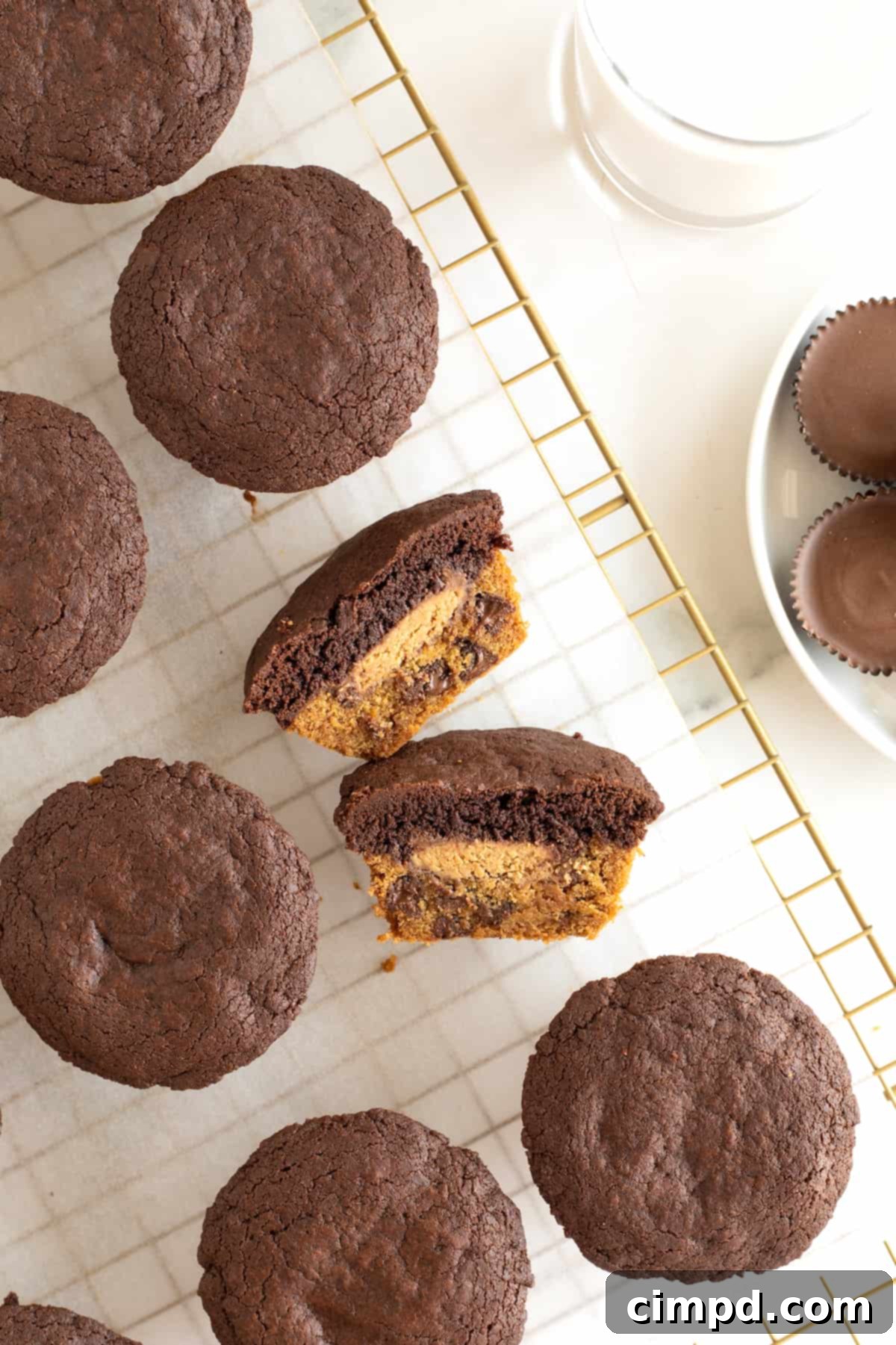 A muffins shaped treat with a cookie bottom, a brownie top and a peanut butter cup in the middle cut in half laying on a parchment lined cooling rack. The rich layers are clearly visible.