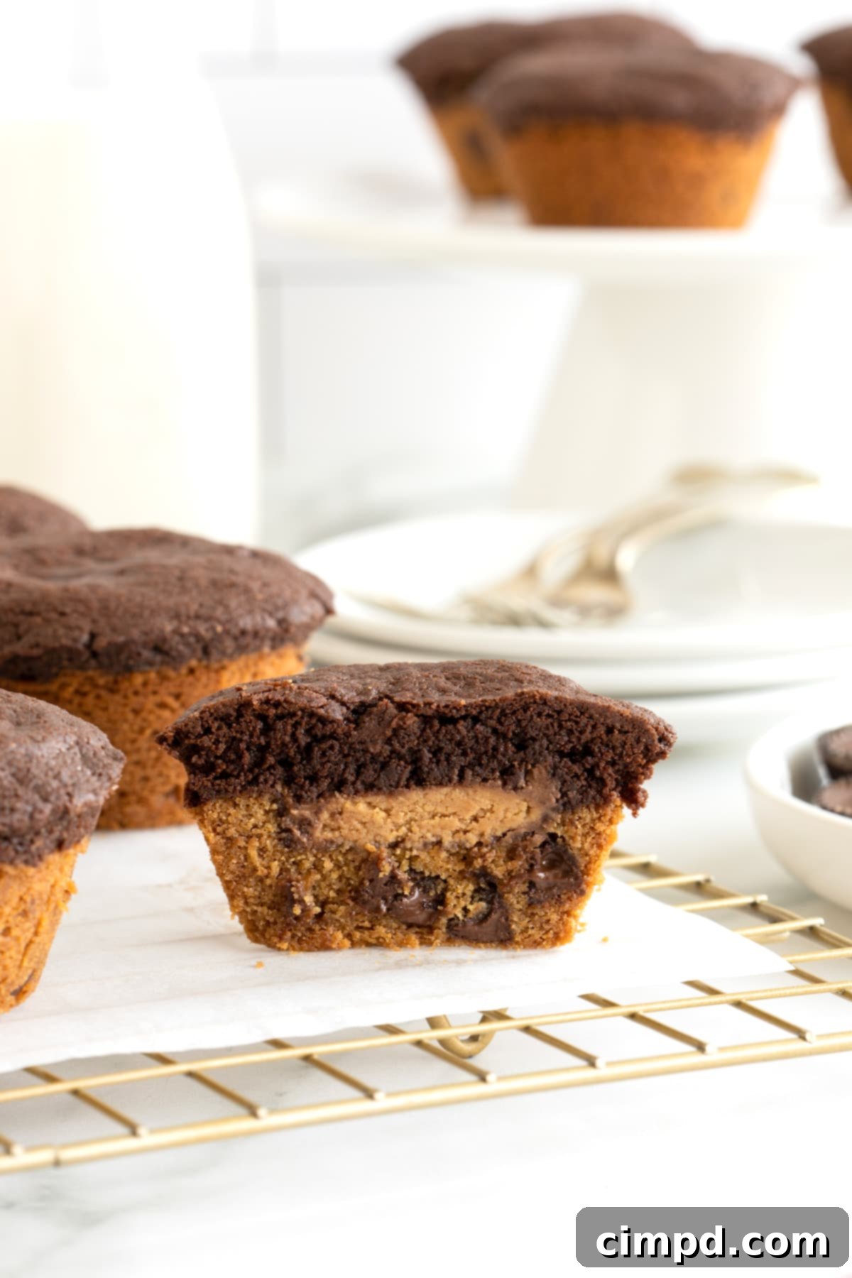 A muffin-shaped treat with a cookie bottom, a brownie top and a peanut butter cup in the middle cut in half laying on a parchment lined cooling rack, illustrating the perfect bake.