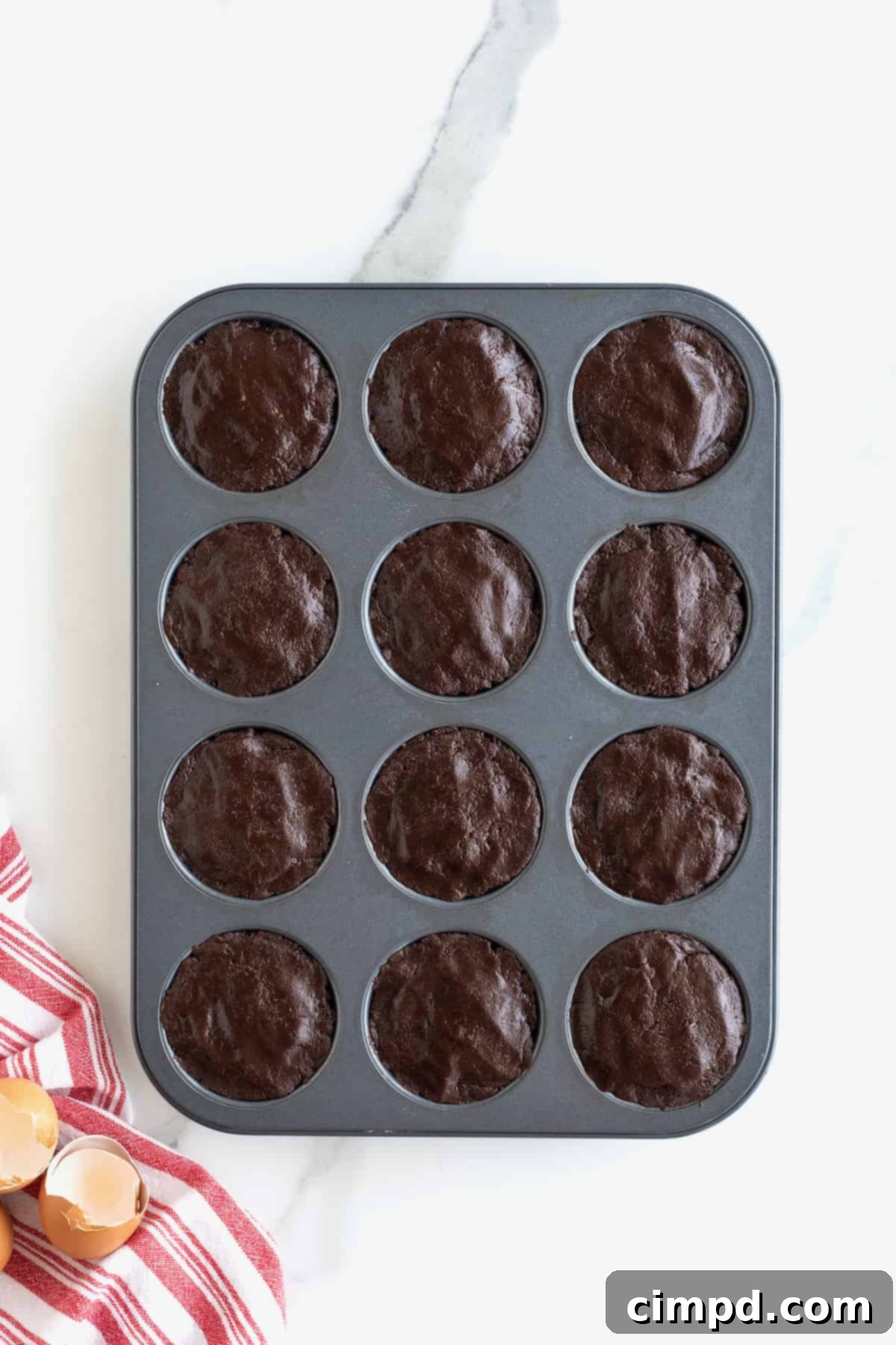 Brownie batter in a dark muffin tin on a white marble counter, neatly spooned over the peanut butter cups.