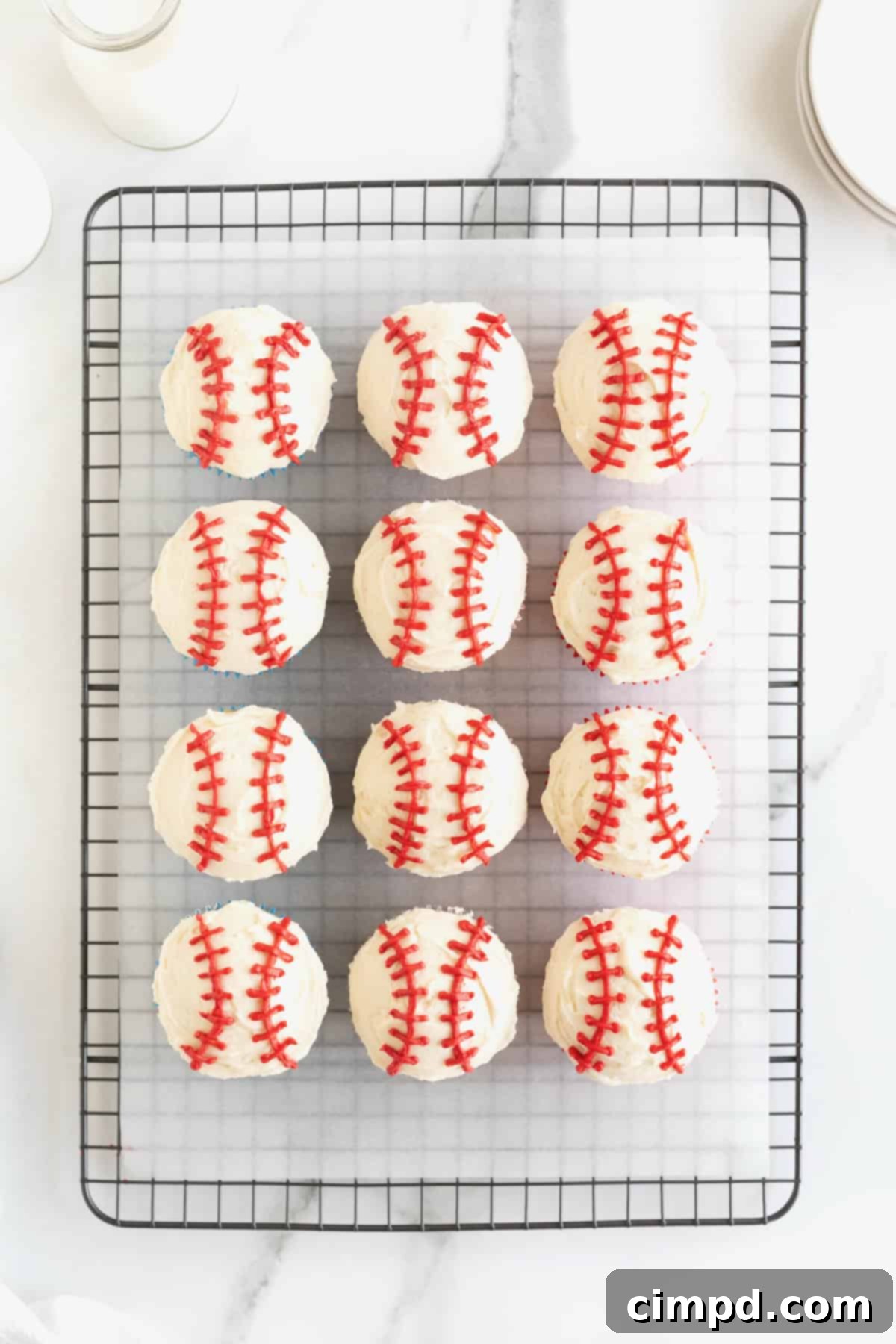 Six beautifully decorated cupcakes on a parchment-lined metal cooling rack, each frosted white with red icing expertly applied to mimic baseball stitching.