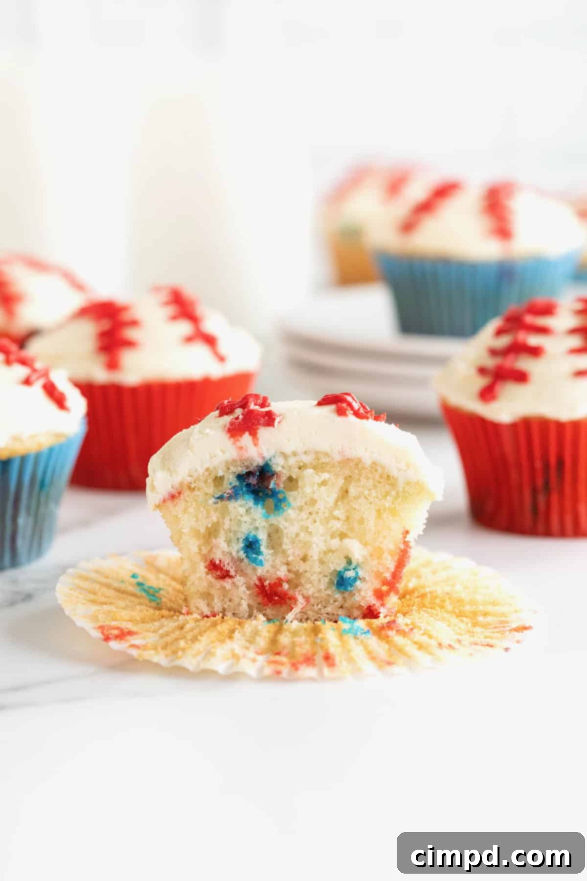 A vibrant confetti cupcake, frosted and decorated to resemble a baseball, with its wrapper partially peeled back to reveal the colorful interior.