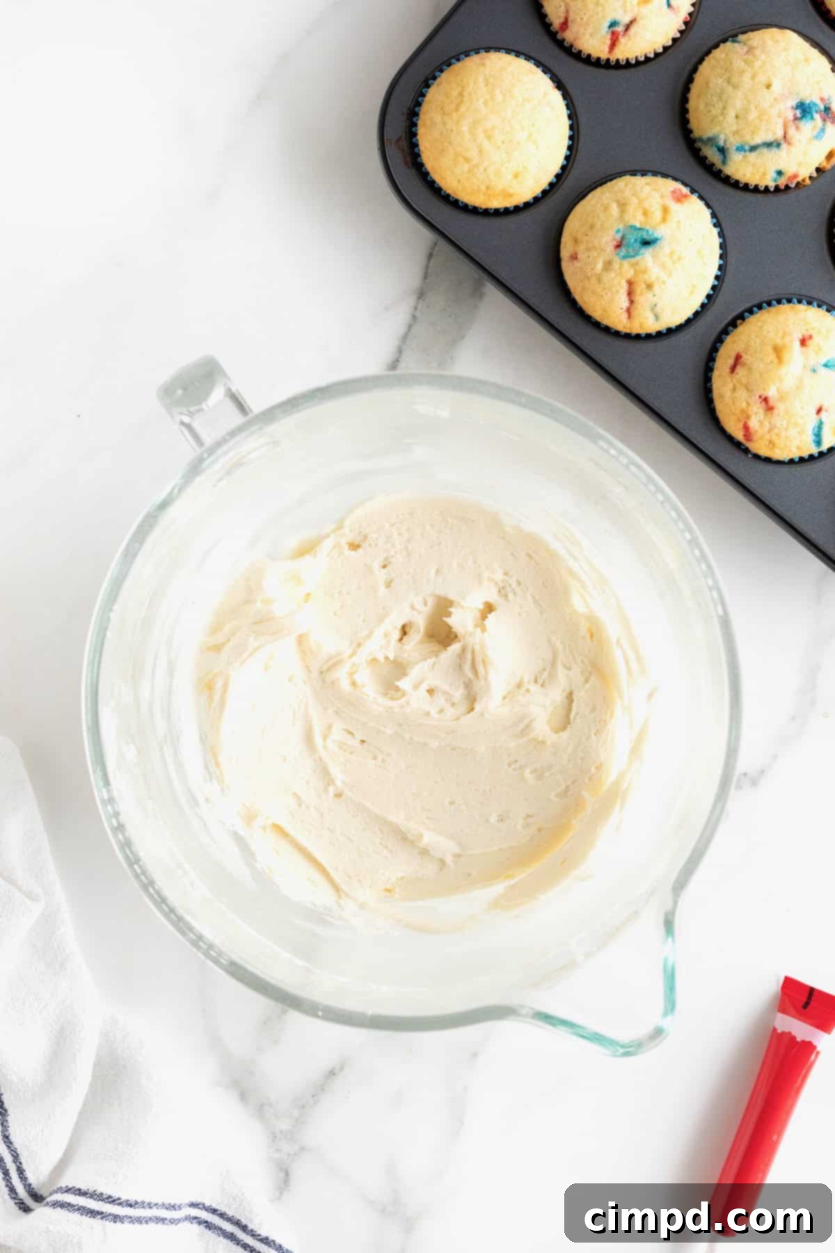 Smooth vanilla frosting in a white glass bowl, positioned beside freshly baked confetti cupcakes in an aluminum muffin pan, ready for assembly.