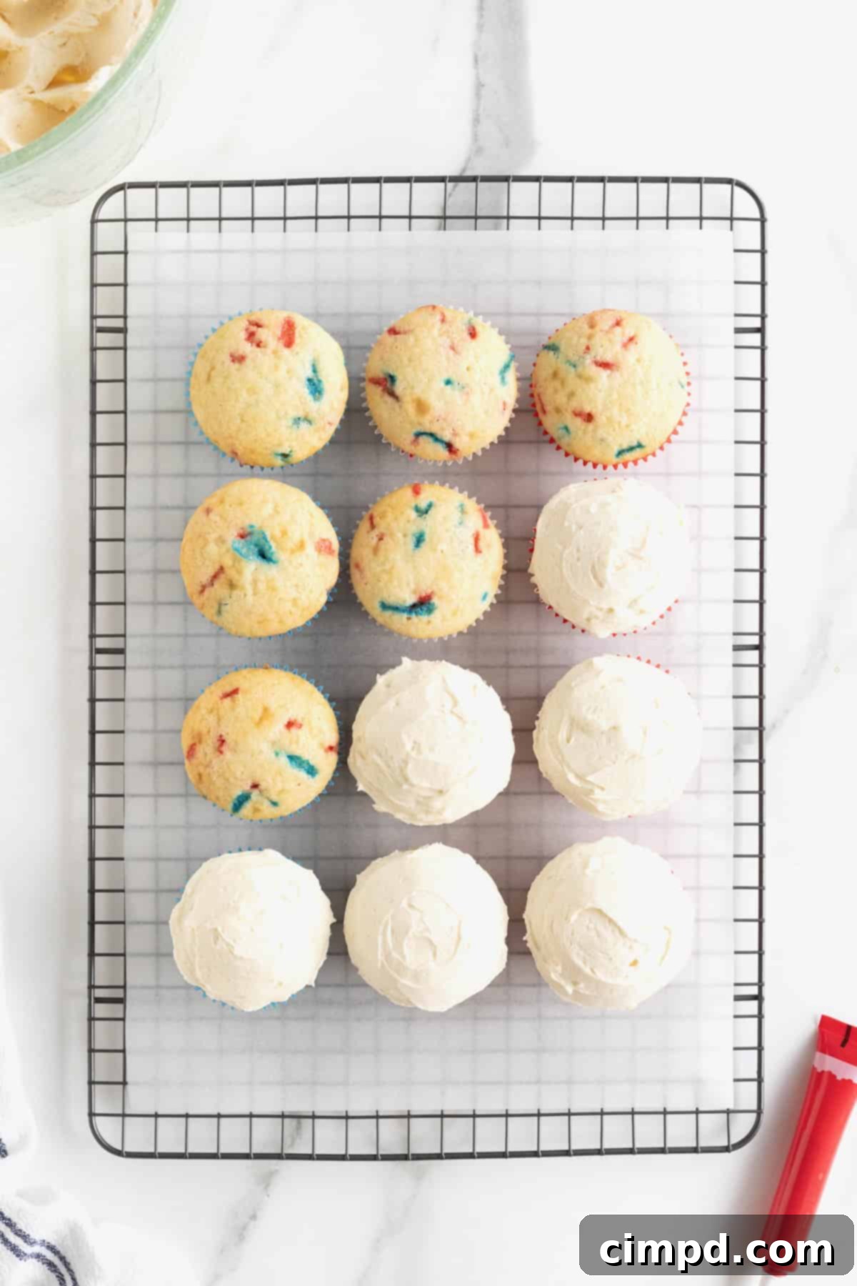 A dozen confetti cupcakes arranged on a parchment-lined metal cooling rack, with six of them already frosted with a smooth layer of white frosting.