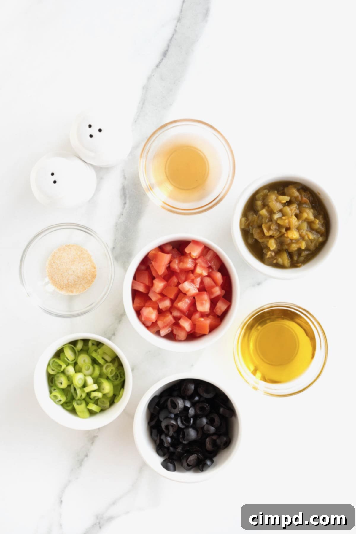 Ingredients for Harlequin Dip arranged in small glass bowls on a white marble counter, including tomatoes, olives, green chilies, and onions.