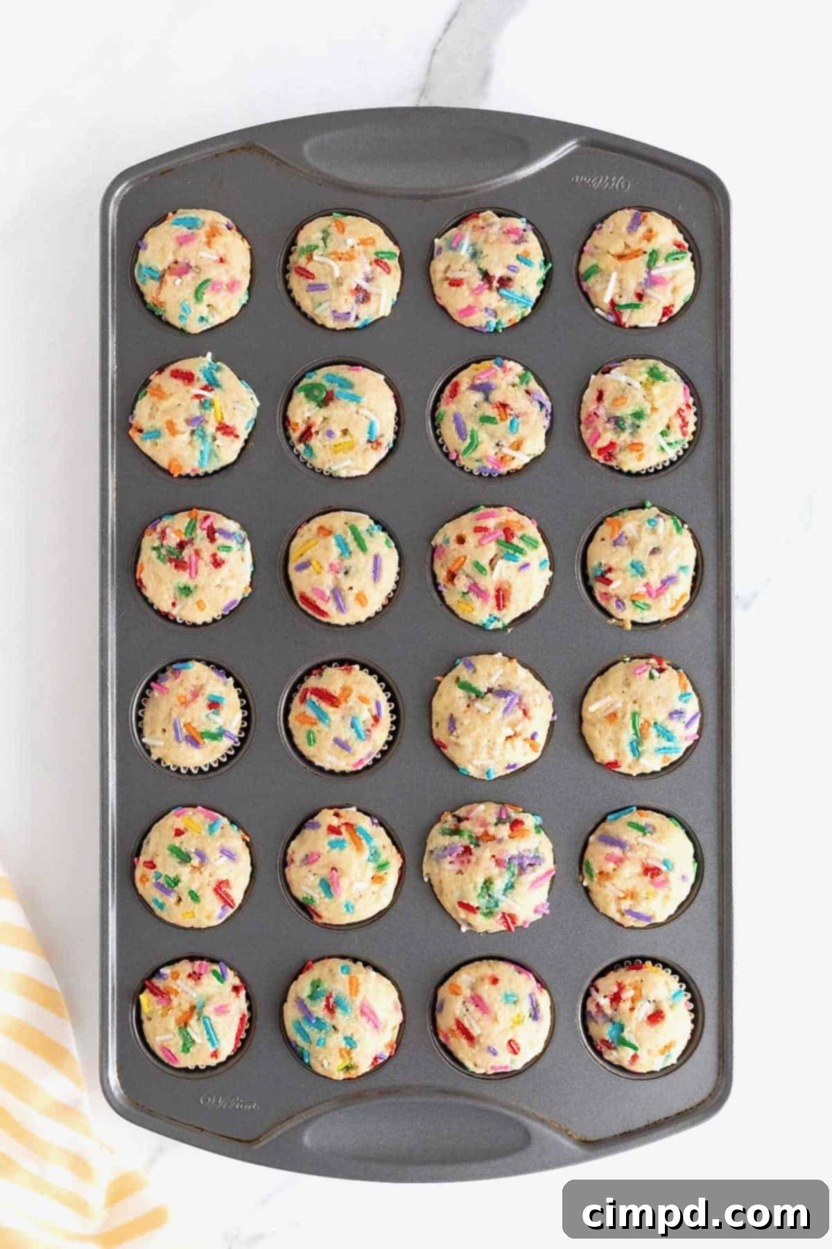 Freshly baked confetti muffins still in a dark aluminum mini muffin pan, their tops golden and speckled with colorful sprinkles.