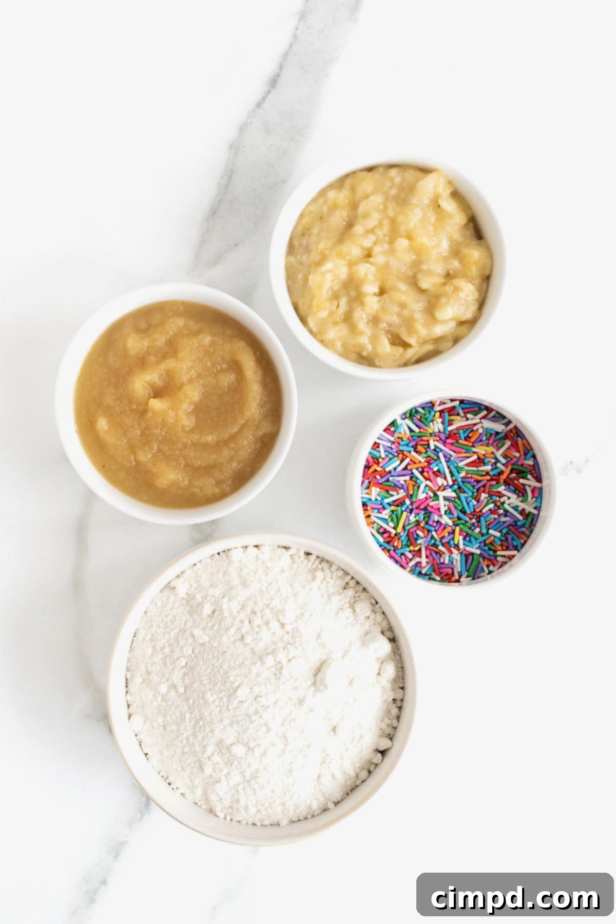 All the simple ingredients for birthday breakfast mini muffins, neatly arranged in small glass dishes on a white marble counter, ready for baking.