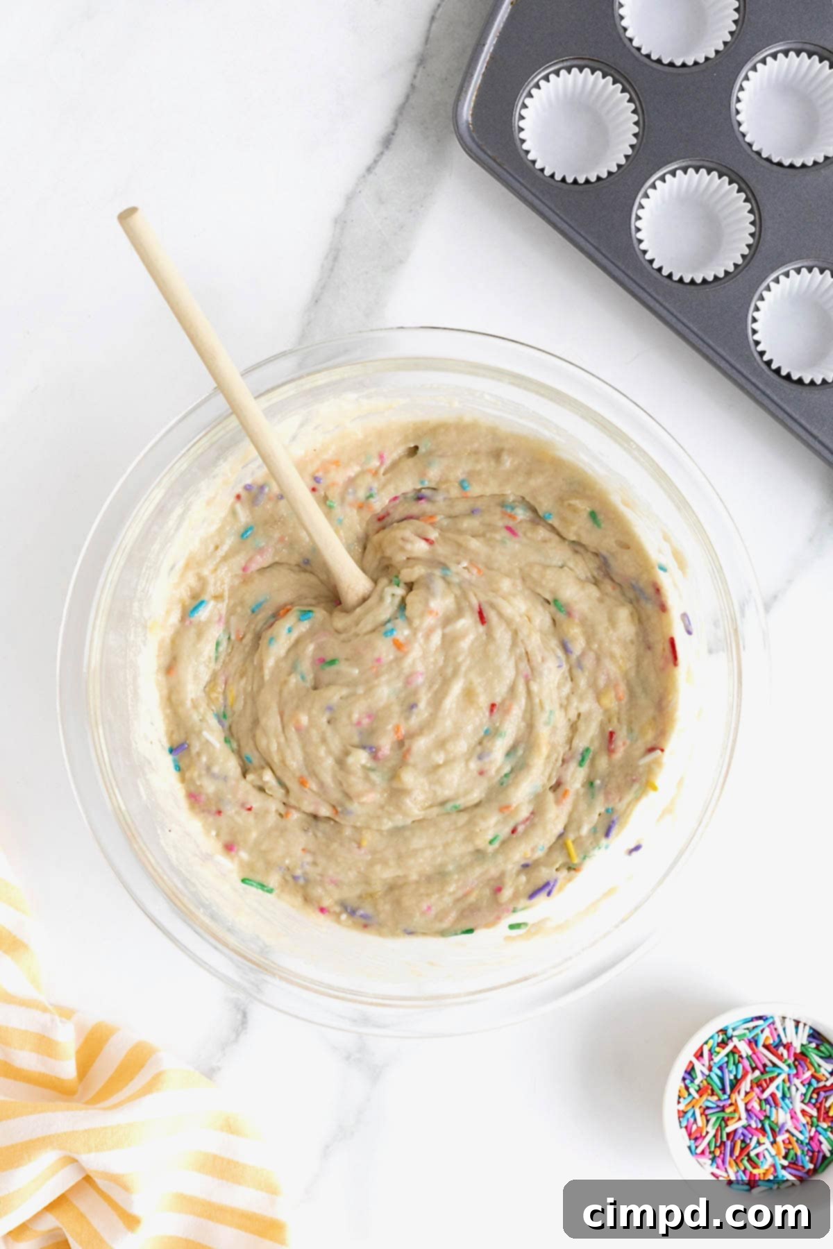 Vibrant rainbow sprinkles being gently folded into the muffin batter in a large clear glass mixing bowl, creating a festive mix.