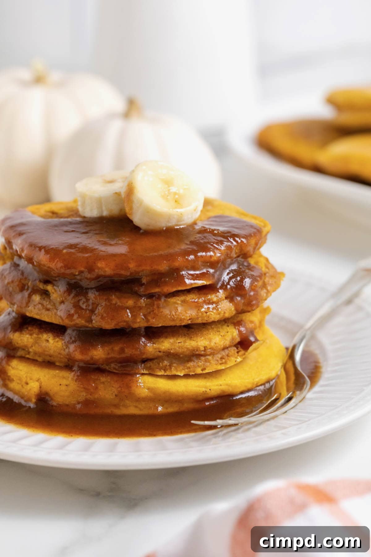 Autumn Pumpkin Banana Pancakes with Pumpkin Spice Drizzle 13 A stack of four banana pumpkin pancakes topped with syrup on a white plate. There are slices of banana on the pancakes and a fork next to them on the plate.