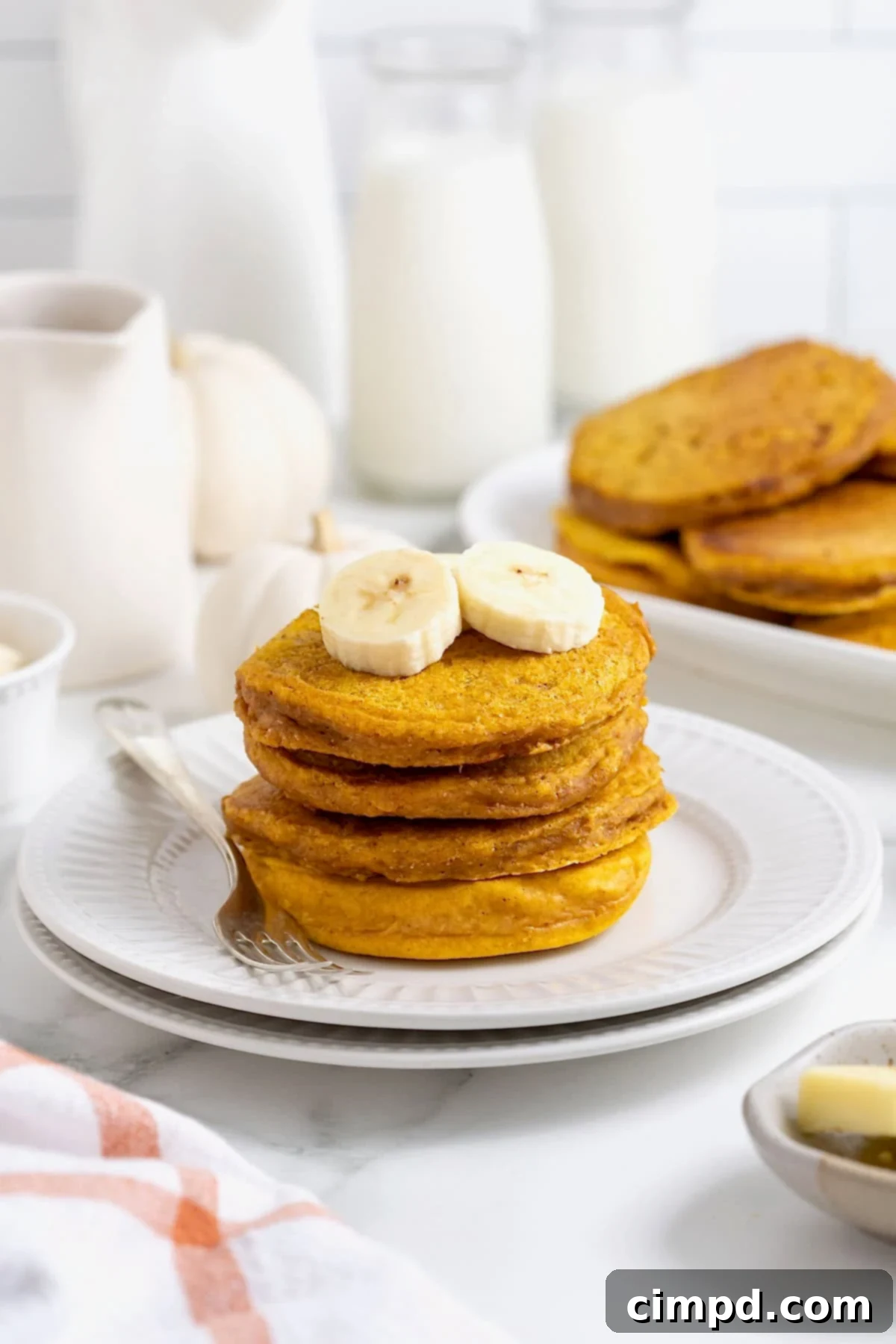 Autumn Pumpkin Banana Pancakes with Pumpkin Spice Drizzle 5 A stack of four pumpkin banana pancakes on a white plate. There are banana slices on top of the pancakes and a fork next to them on the plate.