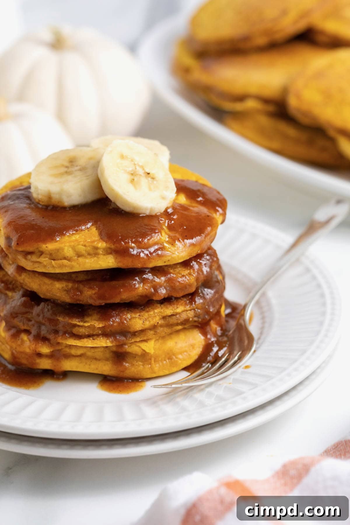 Autumn Pumpkin Banana Pancakes with Pumpkin Spice Drizzle 6 A stack of four banana pumpkin pancakes topped with syrup on a white plate. There are slices of banana on the pancakes and a fork next to them on the plate.
