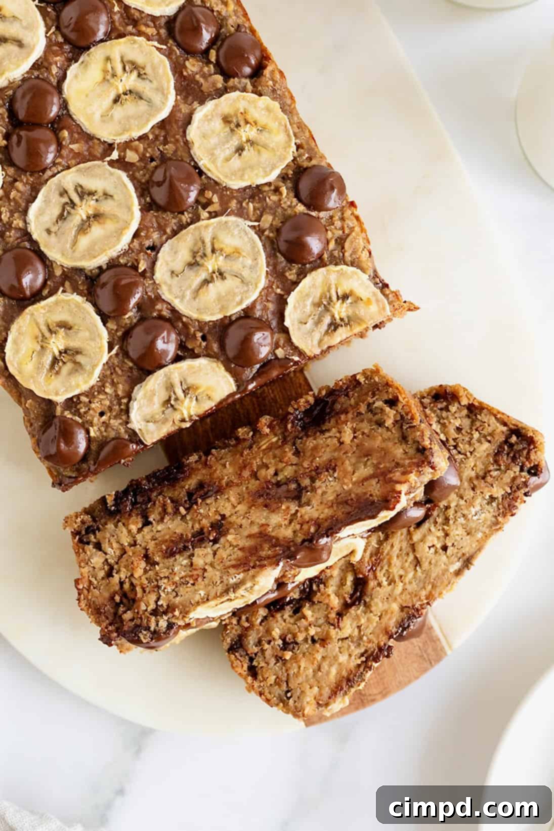 A perfectly baked loaf of flourless chocolate chip banana bread, elegantly garnished with banana slices and a sprinkle of chocolate chips, resting on a pristine white cutting board.