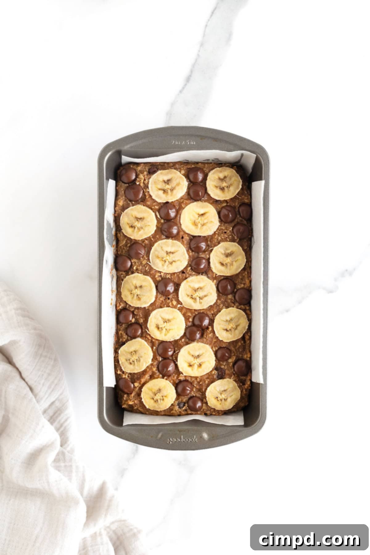 A beautifully baked loaf of chocolate chip banana bread sits cooling in a dark aluminum loaf pan, its golden-brown crust promising deliciousness.