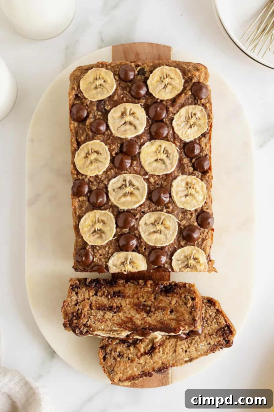 A beautifully presented loaf of banana bread, adorned with fresh banana slices and chocolate chips, rests on a white cutting board, inviting a delicious experience.