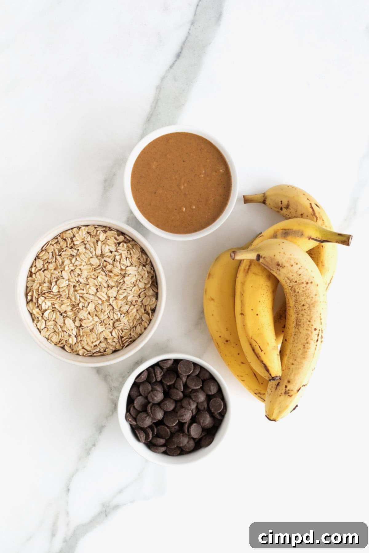The simple quartet of ingredients—ripe bananas, old-fashioned oats, natural peanut butter, and chocolate chips—elegantly arranged in white dishes on a white marble counter, ready to create flourless chocolate chip banana bread.