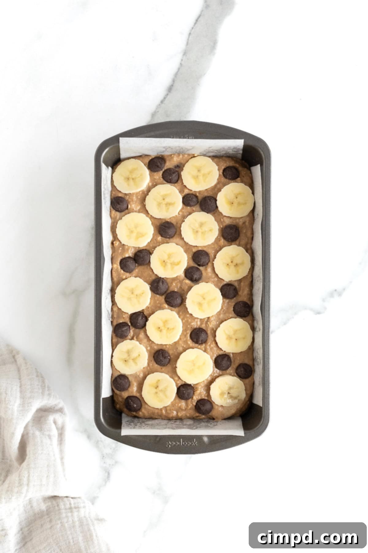 A dark aluminum loaf pan filled with chocolate chip banana bread batter, elegantly garnished with thin banana slices and a scattering of chocolate chips, ready for the oven.