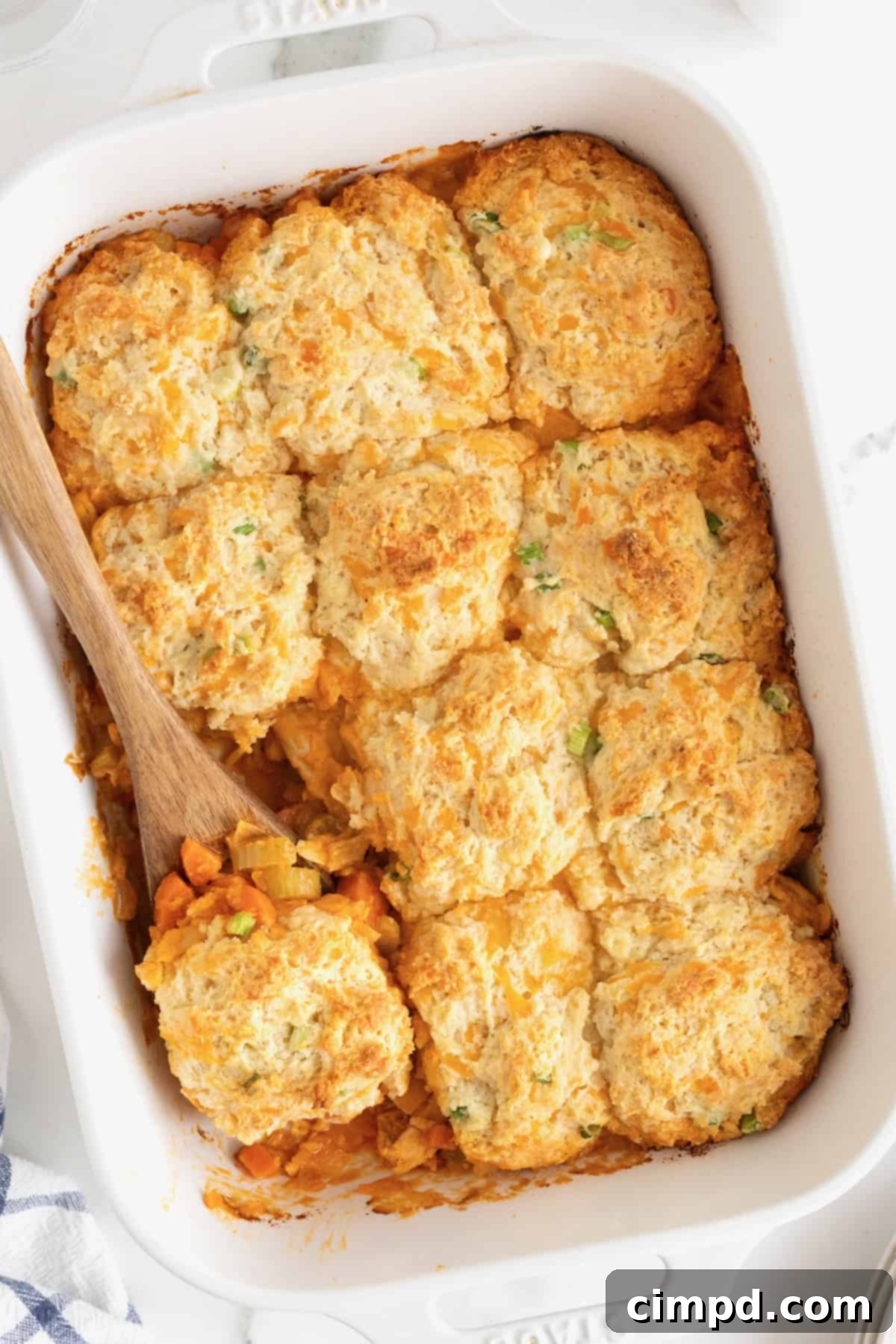 A white casserole dish of buffalo chicken biscuit bake with a wooden spoon in it, showcasing its golden-brown biscuits and bubbly filling.