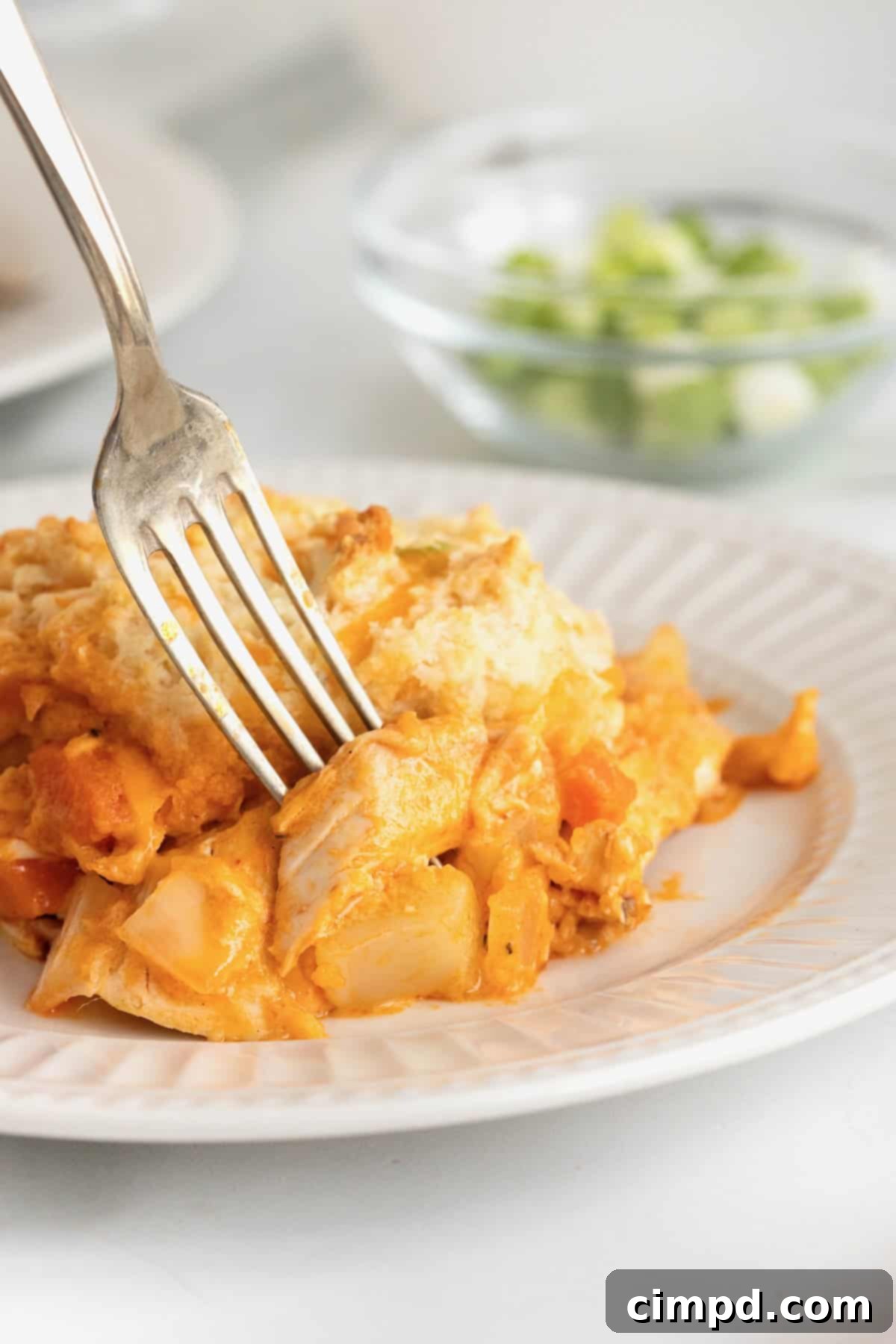 A fork lifting a bite of Buffalo Chicken Biscuit Bake, revealing layers of cheesy biscuit and creamy chicken filling on a white fluted plate.
