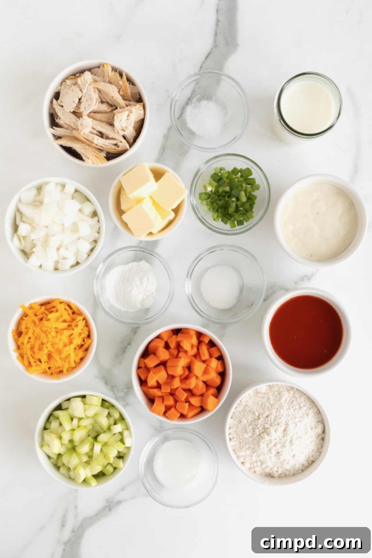An array of fresh ingredients for Buffalo Chicken Biscuit Bake, including chopped vegetables, shredded chicken, cheese, and sauces, neatly arranged in small white and glass dishes on a white marble counter.