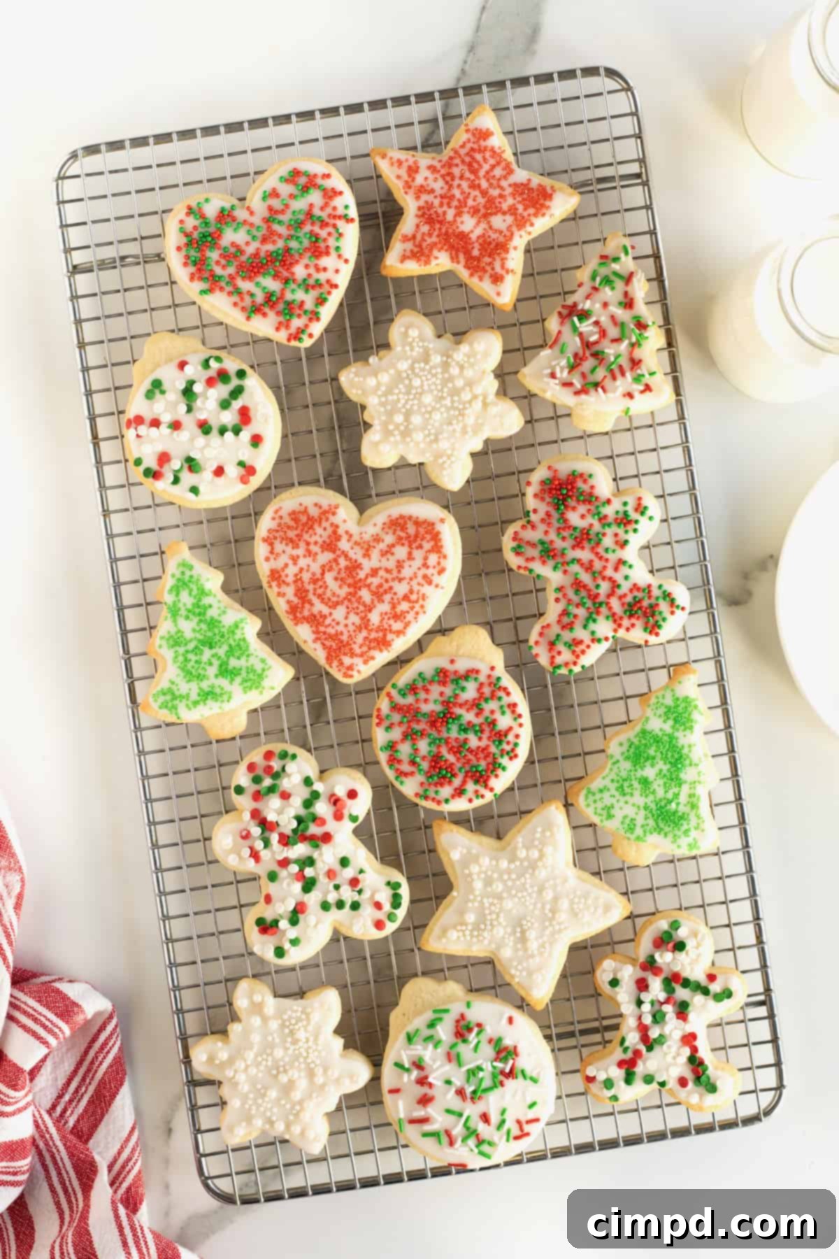 A wire cooling rack filled with sugar cookies shaped like hearts, stars, Christmas trees and gingerbread men decorated with white icing and red and green sprinkles.