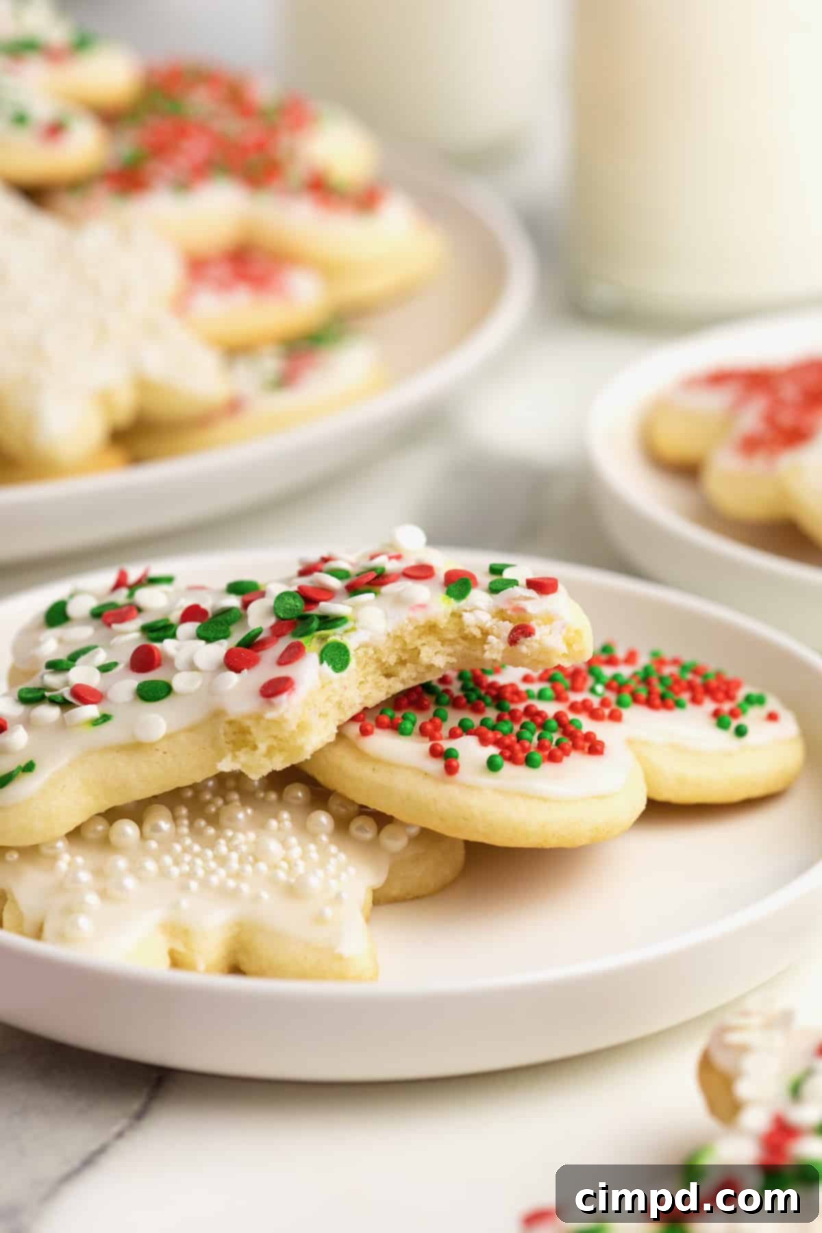 Three frosted Christmas sugar cookies on a white rimmed plate, one snowman, one heart and one star. The star has red and green sprinkles and a bite taken out of it.