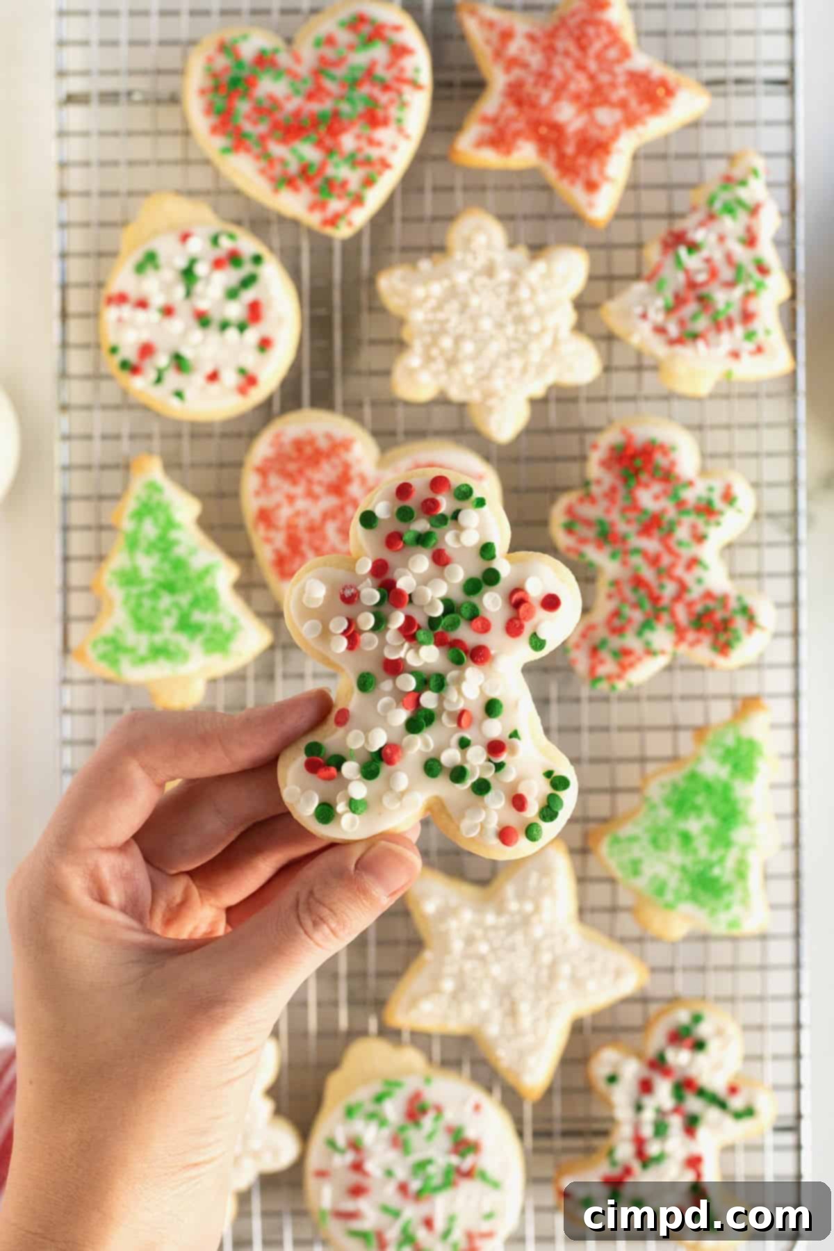 A hand holding a gingerbread man shaped sugar cookie. The cookie has white icing with red, white and green sprinkles. In the background are several more iced holiday sugar cookies on a metal cooling rack.