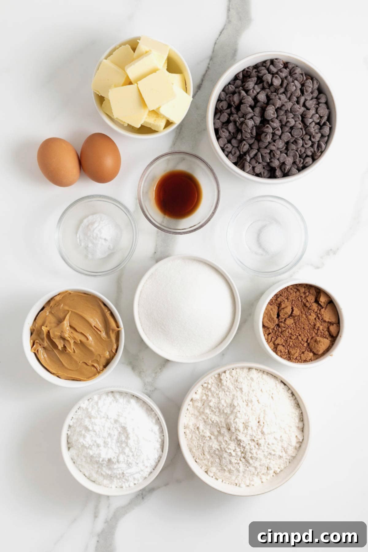Decadent Peanut Butter Chocolate Cookie Tower 7 Various ingredients for chocolate peanut butter layered cookie cake displayed in glass bowls on a white marble surface, including butter, chocolate chips, sugar, cocoa powder, and peanut butter.