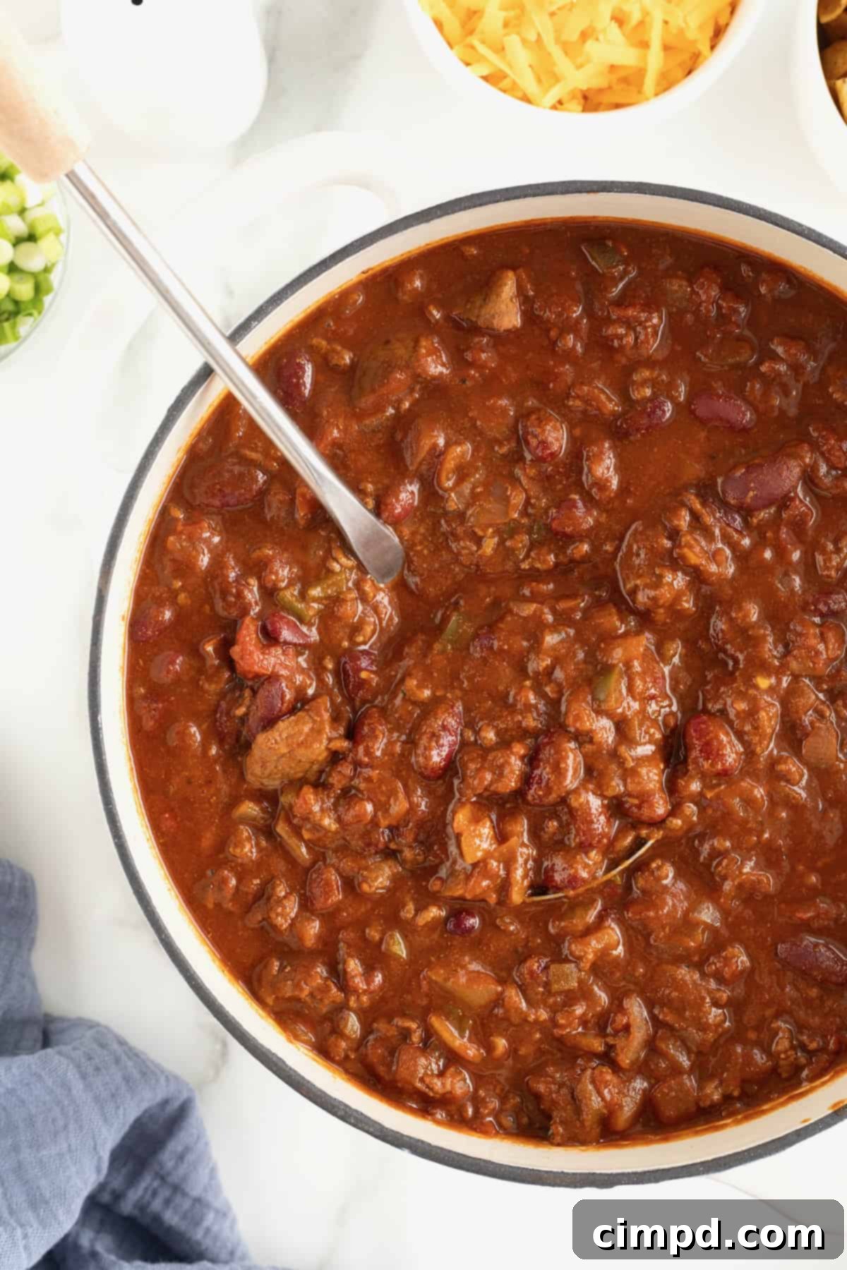 The Gold Standard Chili 11 A large, rustic Dutch oven filled to the brim with a rich, dark, and perfectly cooked chili, with a metal ladle poised inside, set against a pristine white background. This image exudes warmth and hearty flavor.