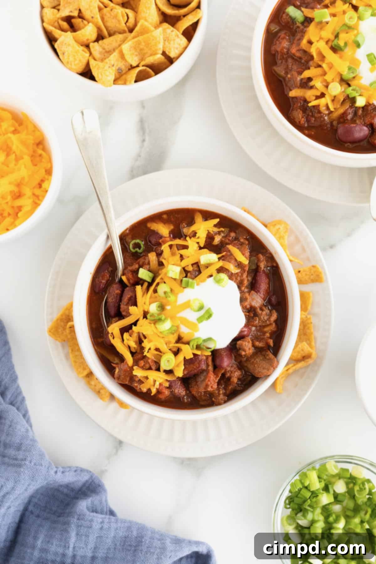 The Gold Standard Chili 3 A pristine white bowl of the best homemade chili, generously topped with shredded cheddar cheese, a dollop of creamy sour cream, and fresh, vibrant sliced green onions. Fritos corn chips are artfully arranged nearby, accompanied by a spoon, ready to be enjoyed.