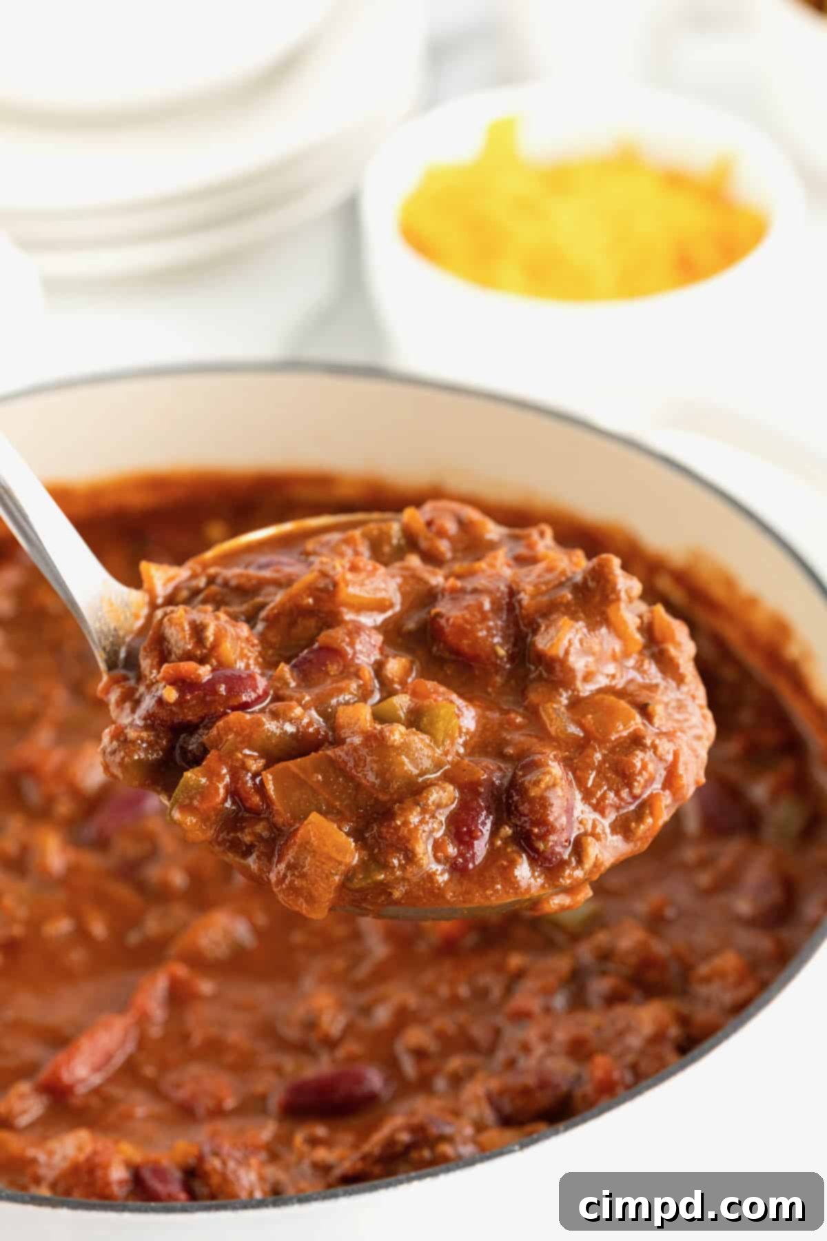 The Gold Standard Chili 4 A hearty Dutch oven filled with a rich, savory homemade chili. A metal ladle hovers, ready to serve, with a small white bowl of shredded cheddar cheese in the soft-focus background, hinting at delicious toppings.