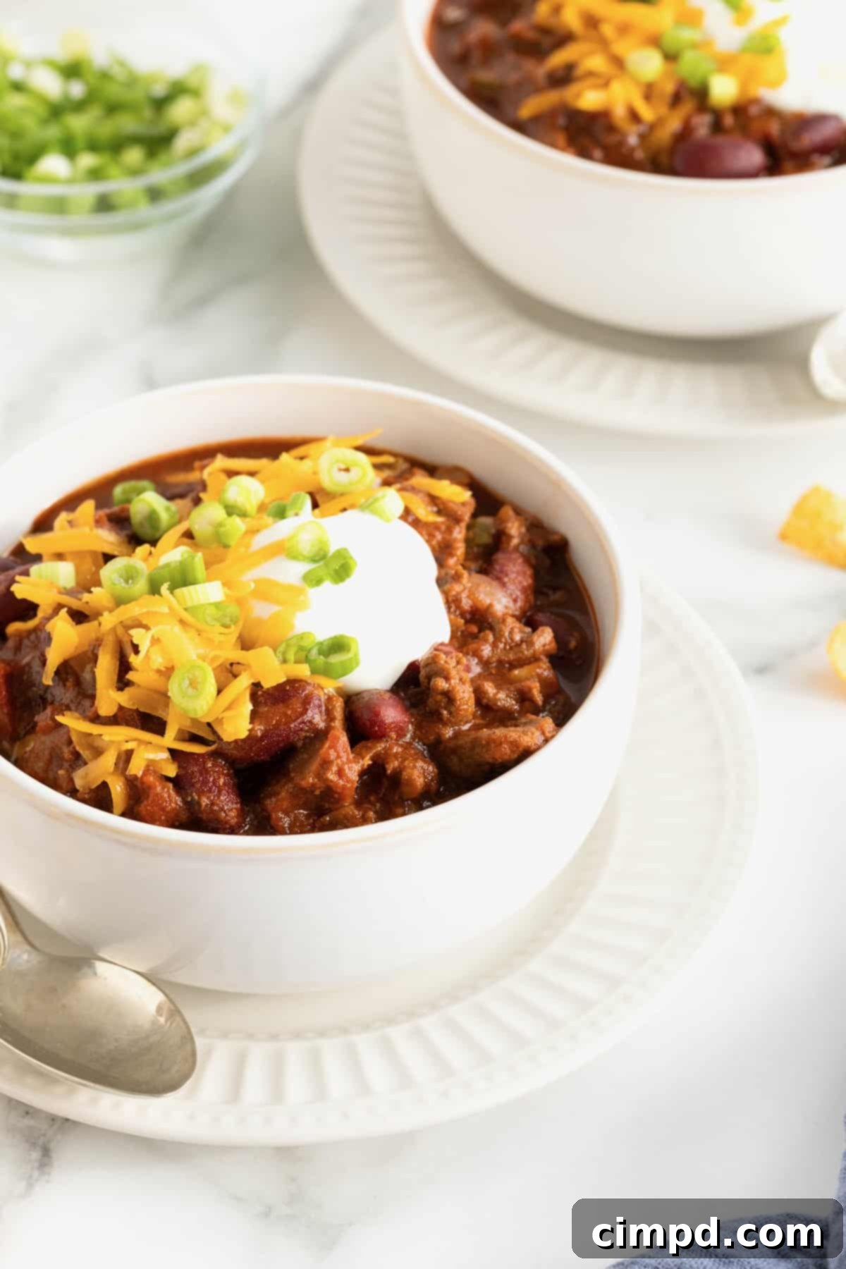 The Gold Standard Chili 5 A pristine white bowl of the best homemade chili, artfully presented on a matching white saucer. The chili is generously adorned with shredded cheddar cheese, a dollop of creamy sour cream, and delicate slices of fresh green onion, making for an appetizing display.