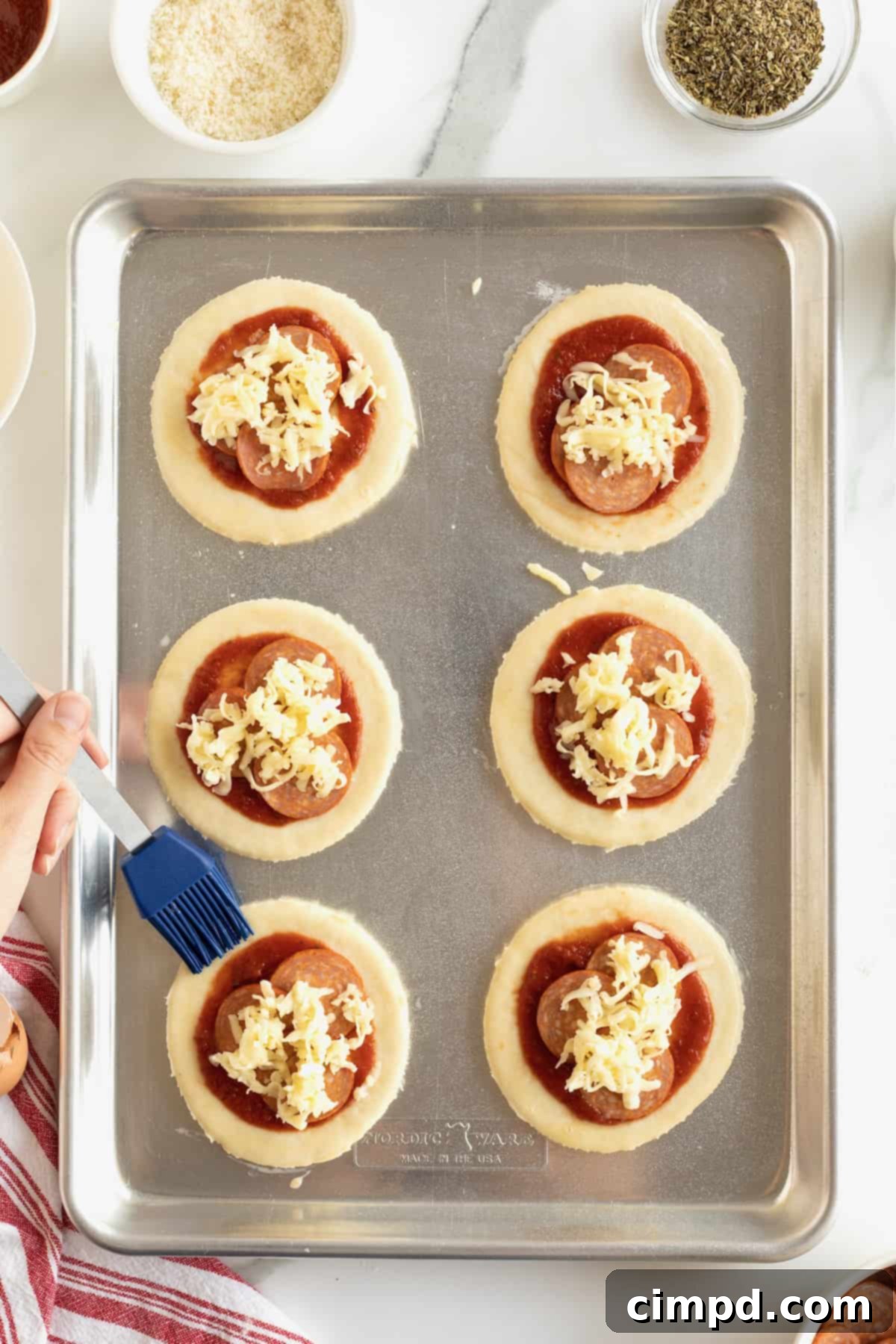Six small circles of dough on an aluminum baking sheet. Each is topped with red tomato sauce, mini pepperonis and cheese. The edges are being brushed with a blue silicon food brush.