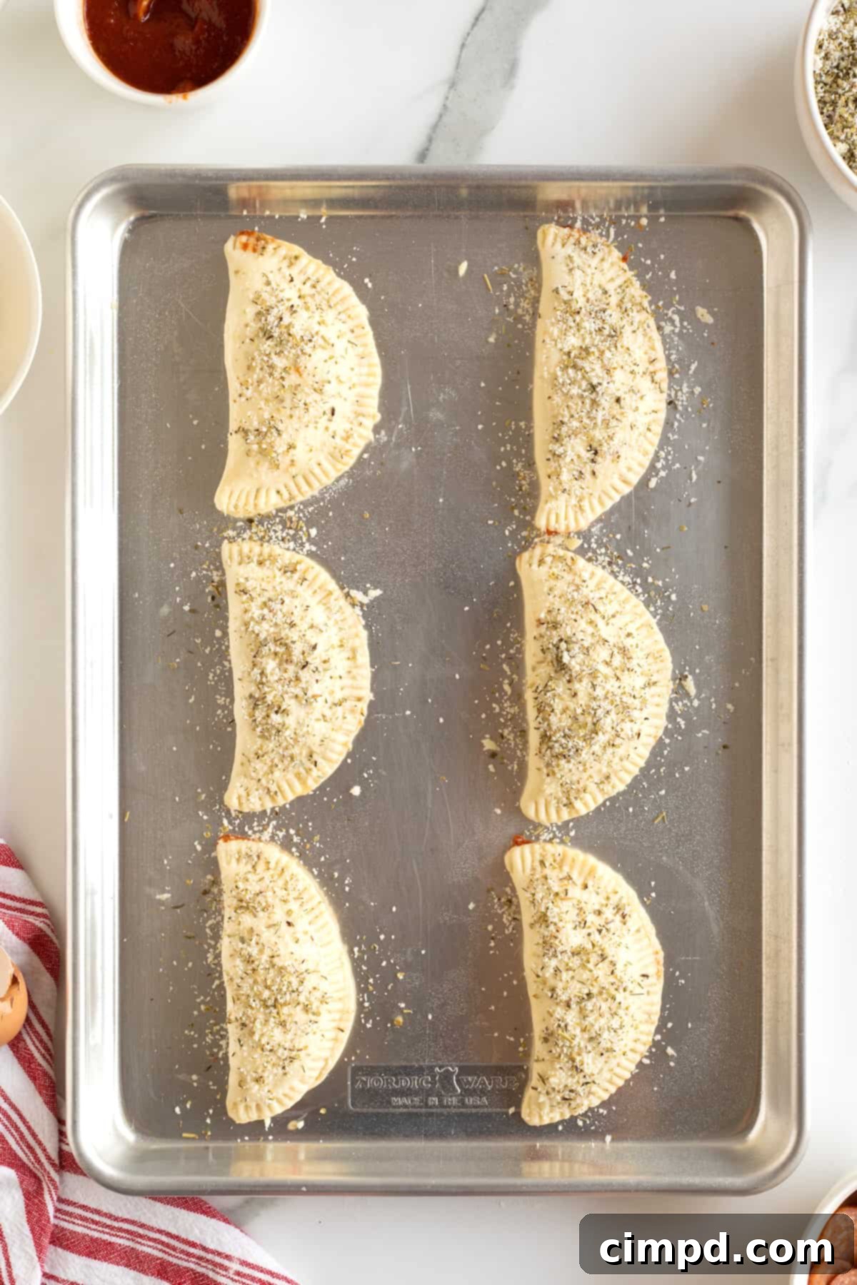 Six unbaked  mini calzones on an aluminum baking sheet.
